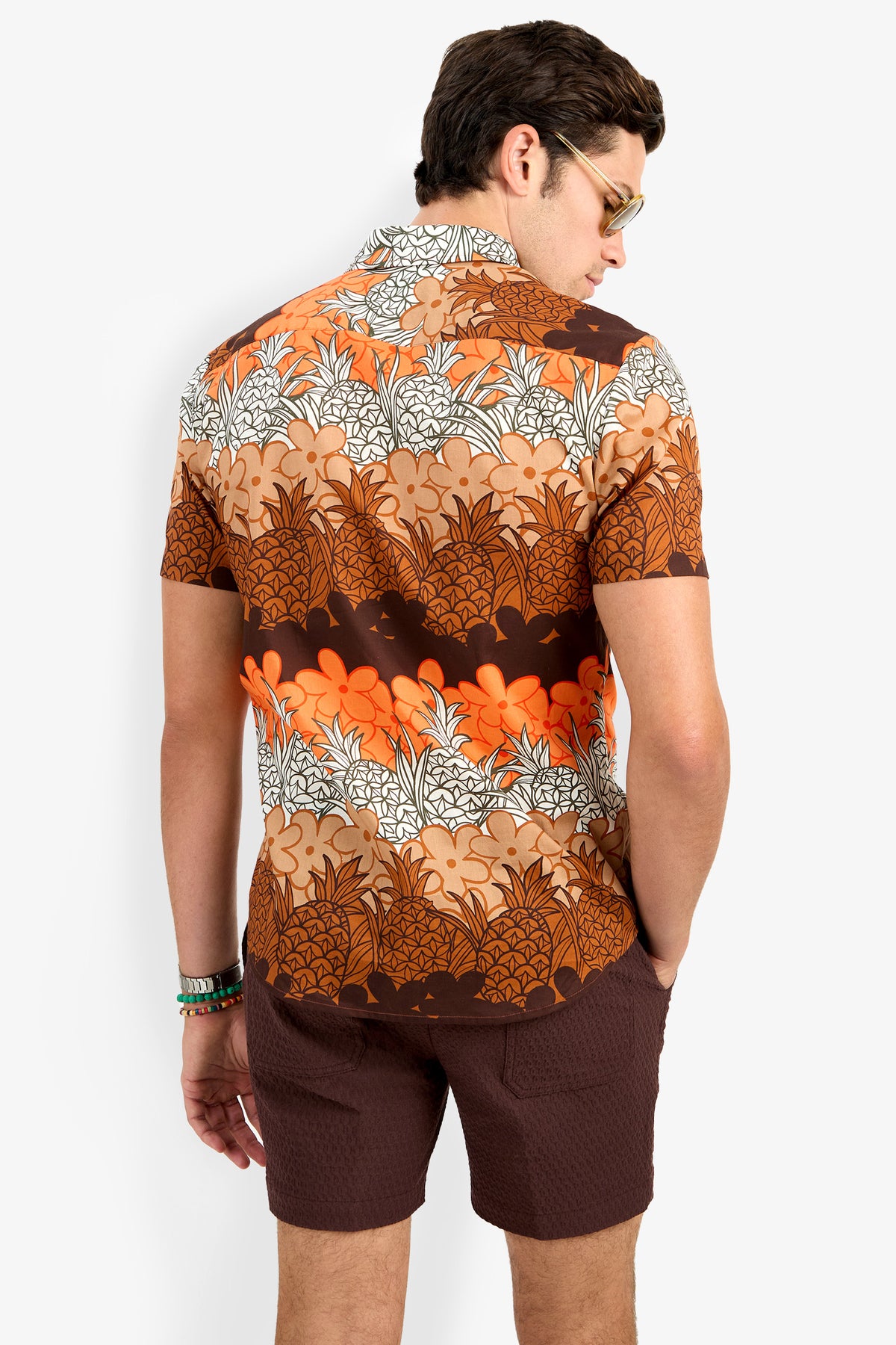 A man with dark hair, sunglasses, and a bracelet on his left wrist stands with his back to the camera, wearing the Slim Jim Shirt—a short-sleeve brown, orange, and white floral-pattern shirt—and brown shorts.