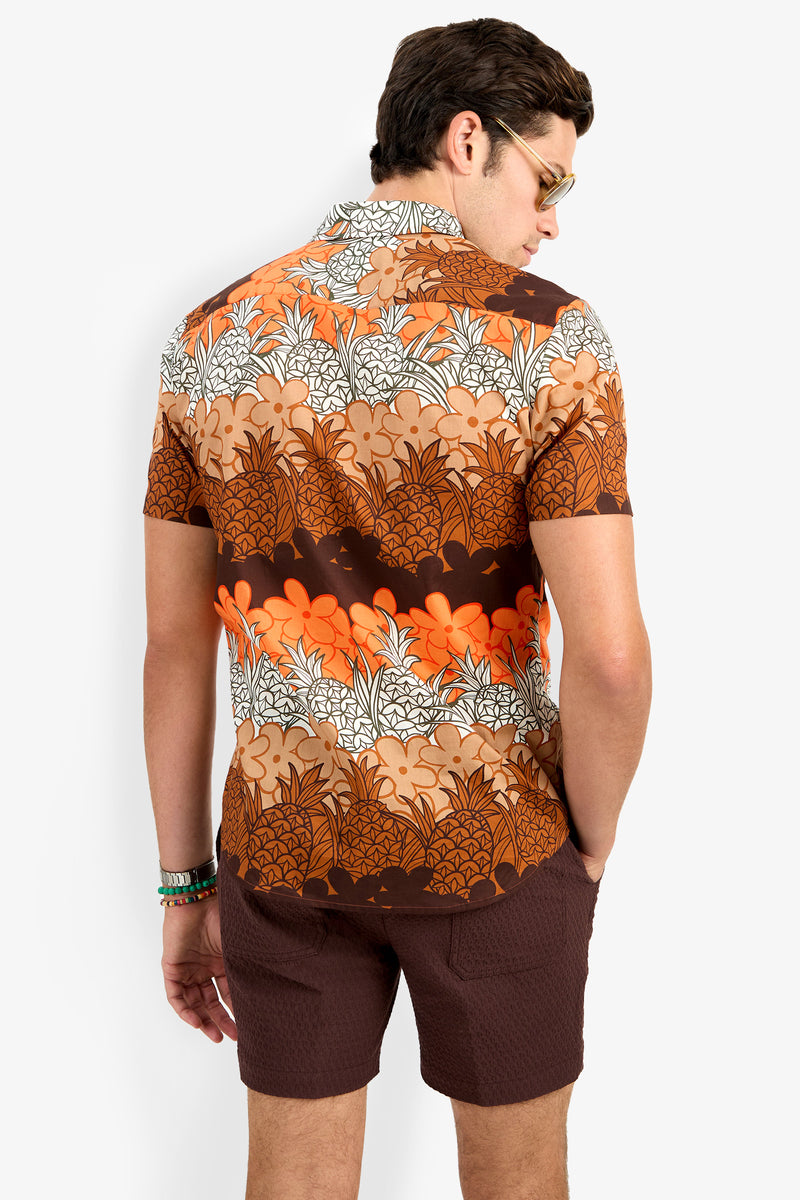 A man with dark hair, sunglasses, and a bracelet on his left wrist stands with his back to the camera, wearing the Slim Jim Shirt—a short-sleeve brown, orange, and white floral-pattern shirt—and brown shorts.