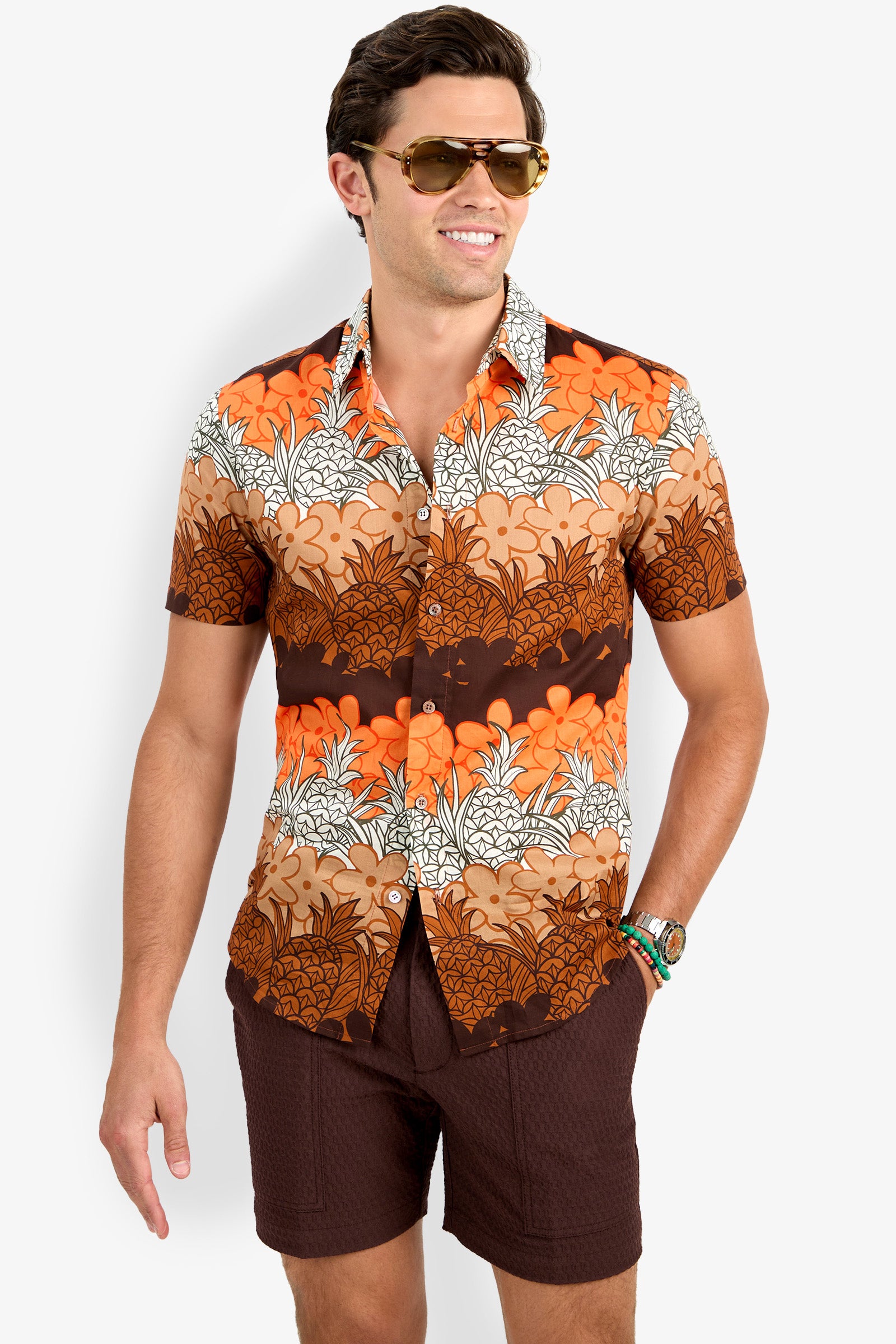 A man stands smiling against a white background, wearing the Slim Jim Shirt—a brown and orange floral short-sleeve style—paired with sunglasses and brown shorts, with one hand in his pocket.