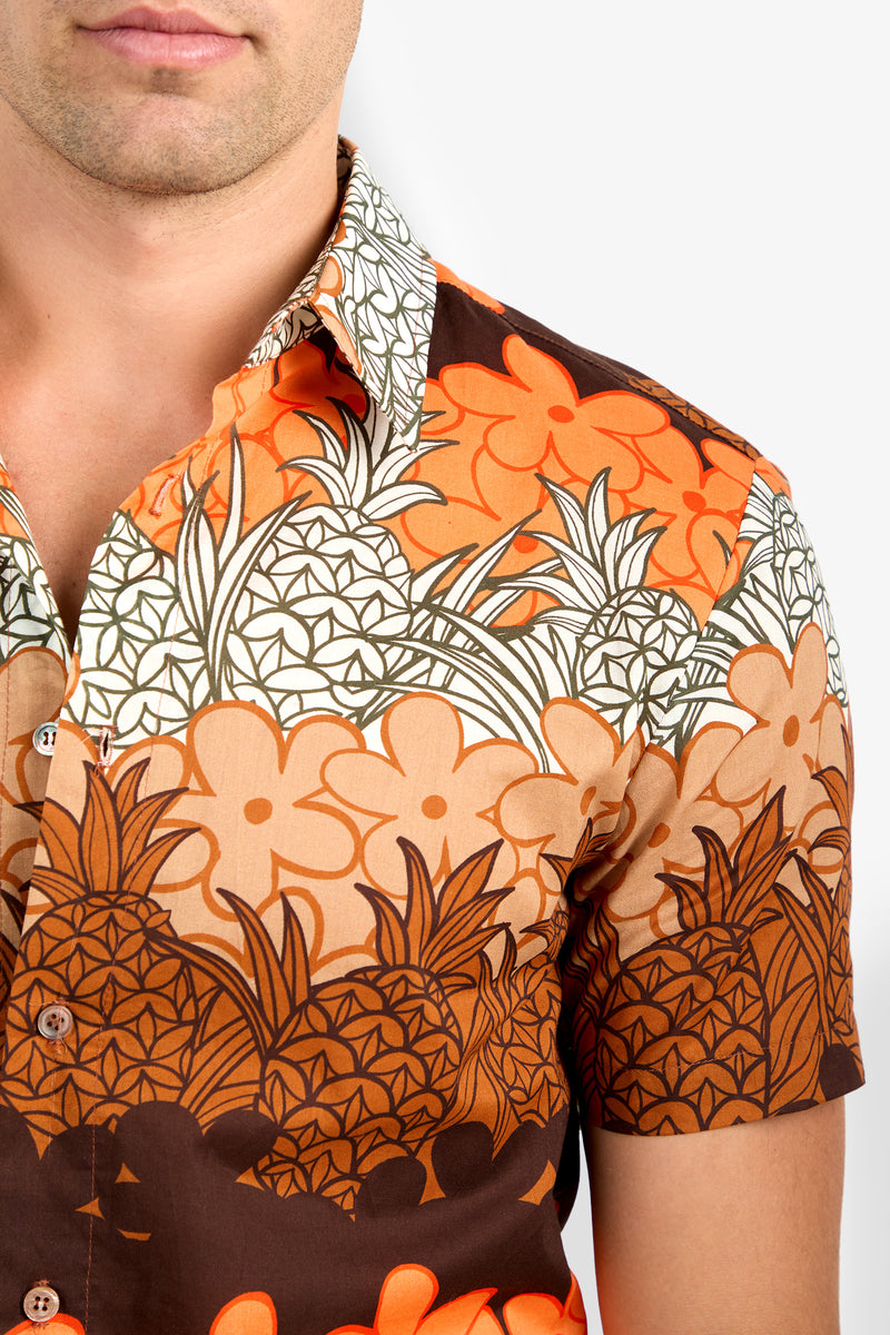 A close-up of a man wearing the Slim Jim Shirt, a short-sleeve button-up with a bold orange, brown, and cream floral and pineapple pattern. Only the lower half of his face is visible.