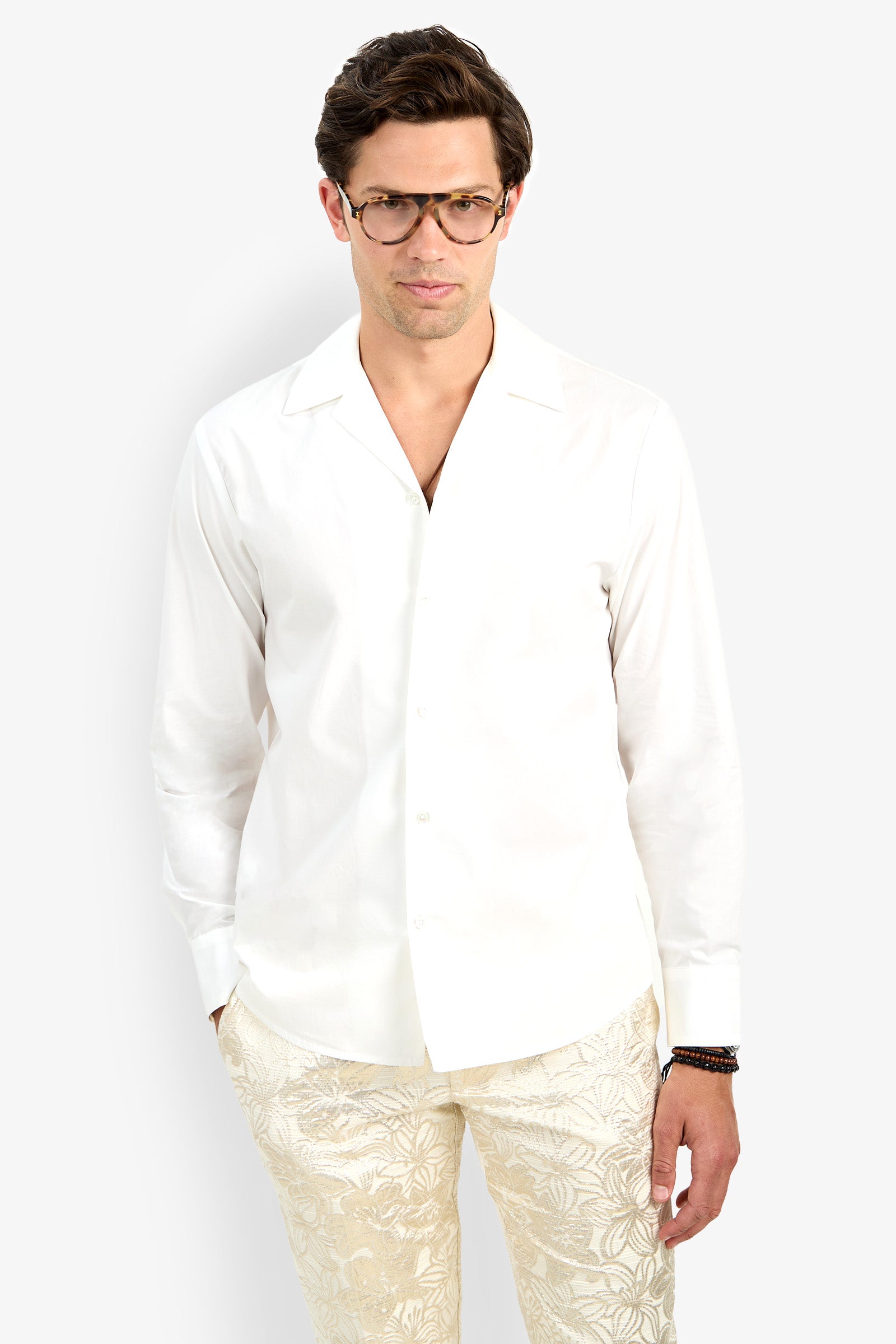 A man wearing glasses and the Able Shirt—a white, long-sleeve button-up—paired with light leaf-patterned pants stands against a plain white background with his hands in his pockets.