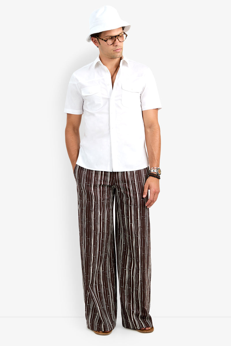 A man stands facing forward with his hands in his pockets, wearing the Shelton Short Sleeve Shirt in white, a white hat, glasses, and brown wide-leg pants with vertical white stripes.