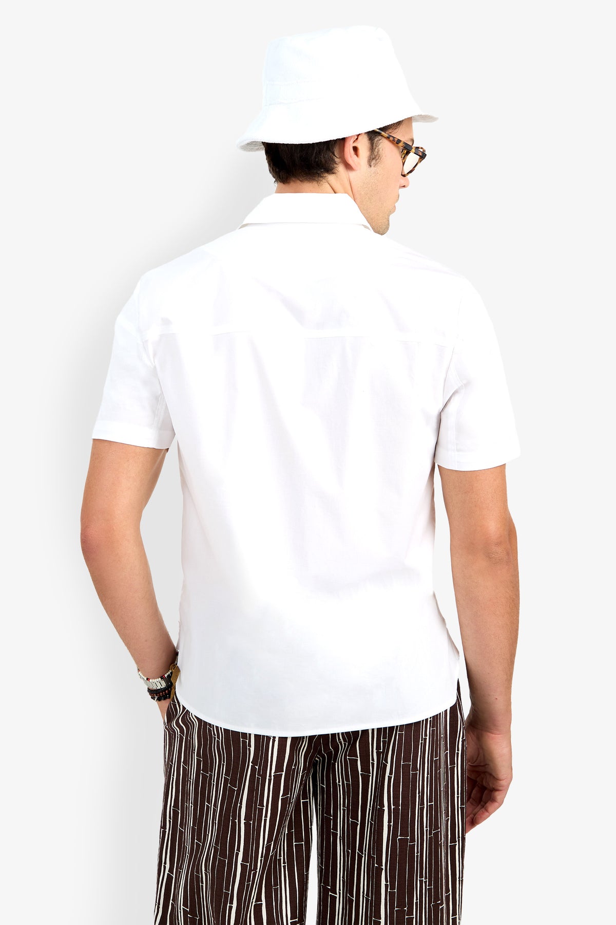 Man facing away, wearing the Shelton Short Sleeve Shirt, a white bucket hat, sunglasses, and brown striped pants against a plain light background.