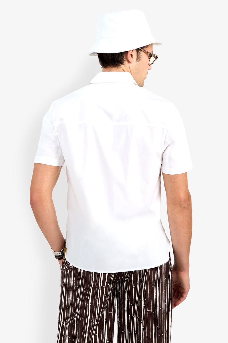 Man facing away, wearing the Shelton Short Sleeve Shirt, a white bucket hat, sunglasses, and brown striped pants against a plain light background.
