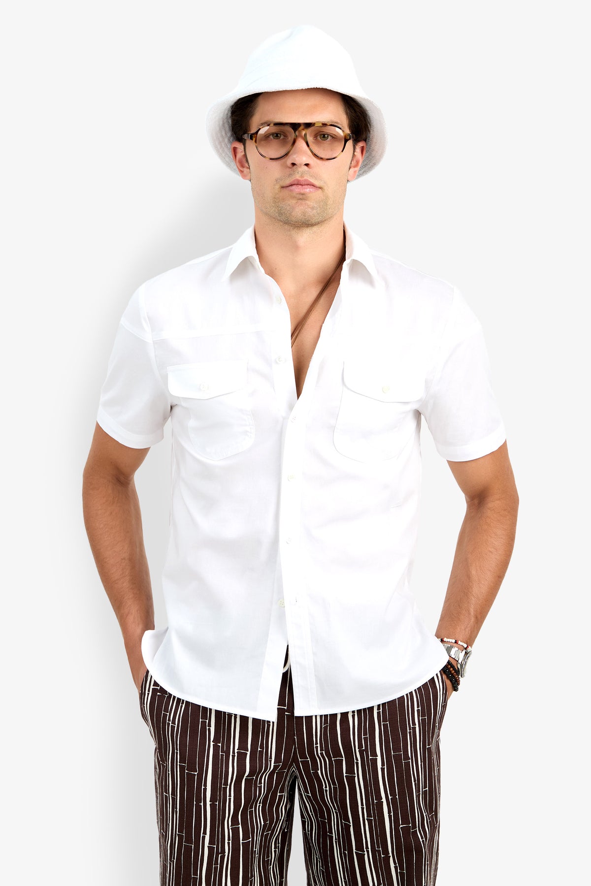 A man stands with his hands in his pockets, wearing glasses, a white hat, patterned brown and white pants, and the Shelton Short Sleeve Shirt against a plain light background.