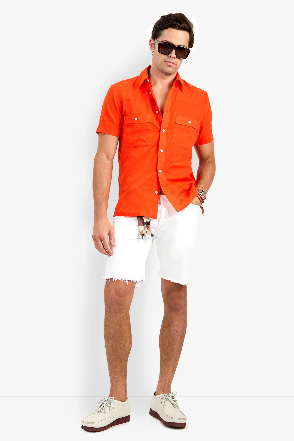A man models the Will Shirt, a bright orange short-sleeve button-up, paired with white frayed shorts, beige shoes, and black sunglasses. He stands relaxed against a plain white background with one hand in his pocket.