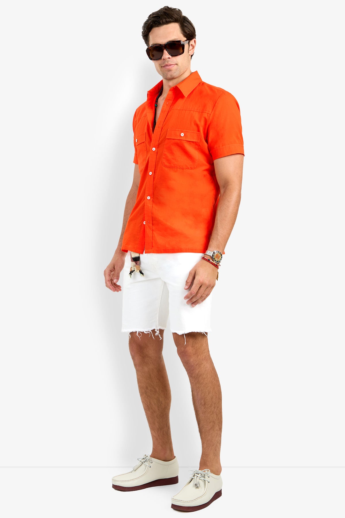 A man models the Will Shirt, a bright orange short-sleeve button-up, styled with white frayed-hem shorts, white shoes, sunglasses, and a watch, posing confidently against a plain white background.