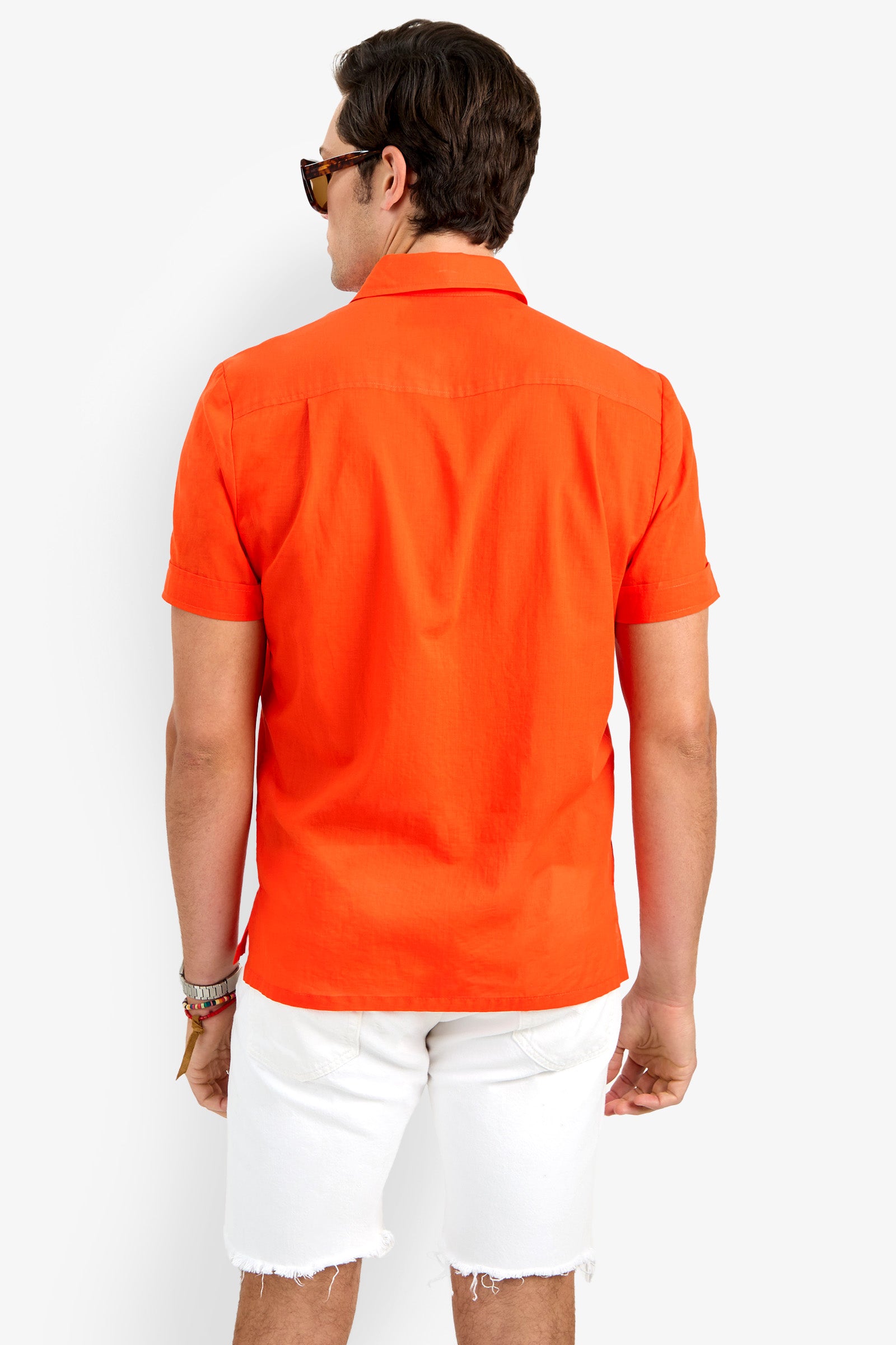 A man wearing sunglasses and the orange Will Shirt, a short-sleeve button-up, with white frayed shorts stands facing away from the camera against a plain white background.