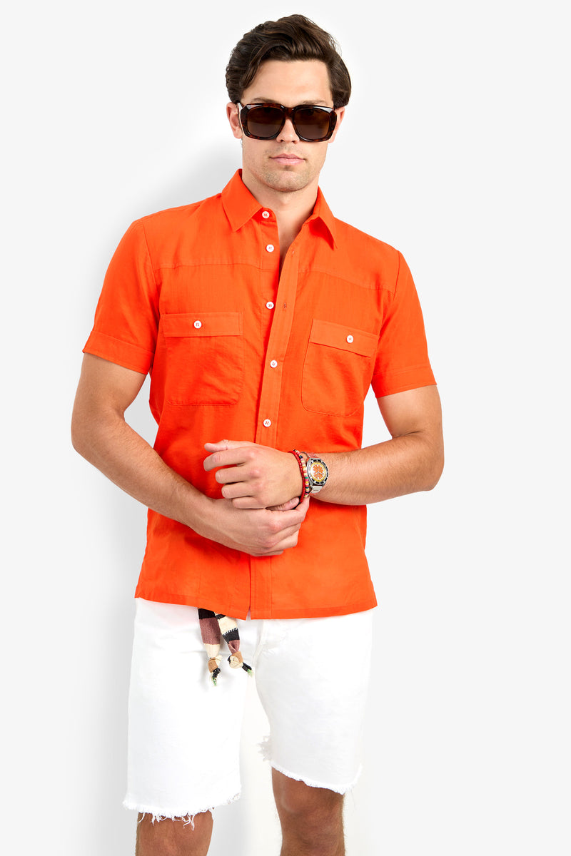 A man in dark sunglasses and the Will Shirt, an orange short-sleeve button-up with front pockets, paired with white frayed shorts stands against a plain white background. He accessorizes with a watch and bracelets on his left wrist.