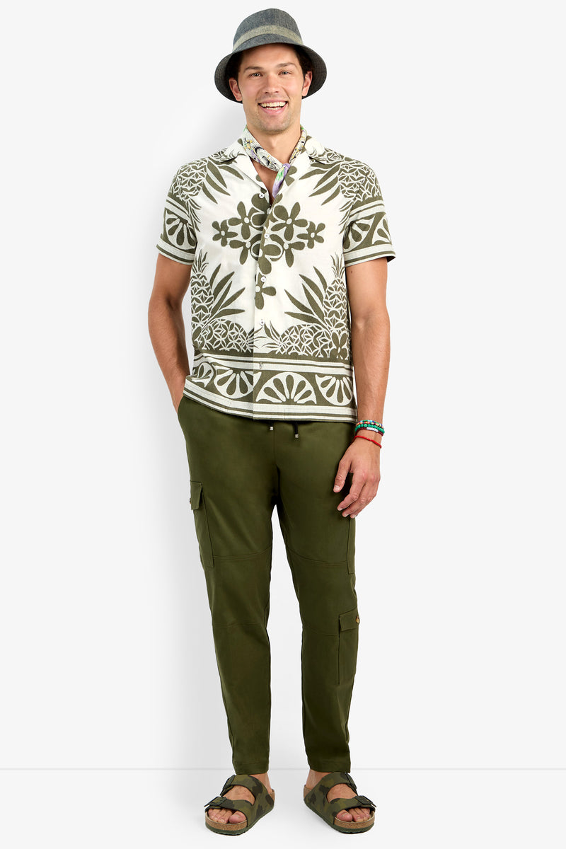 A smiling man wearing a gray hat, Felix Shirt, olive green cargo pants, sandals, and colorful bracelets stands against a white background.