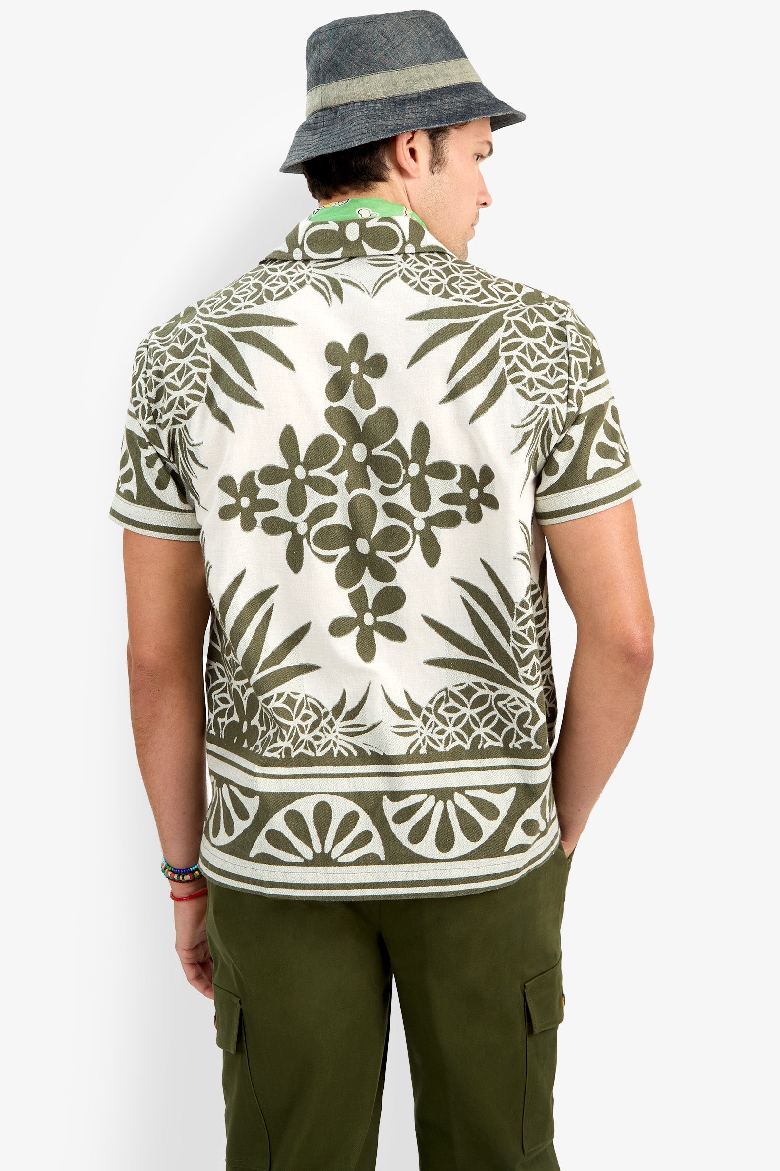 A person seen from behind wears a gray bucket hat, olive green cargo pants, and the Felix Shirt, which features a short-sleeve design with an olive green and white floral pattern.