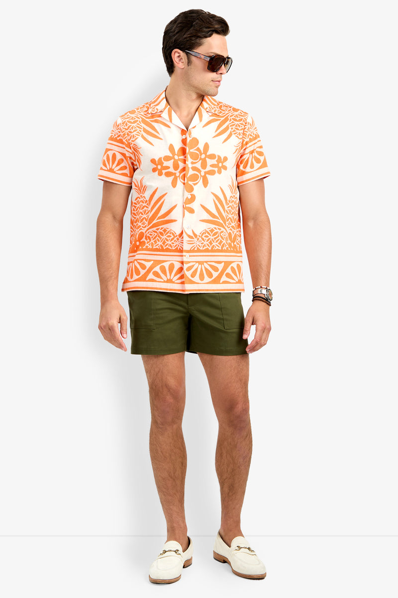 A man wearing sunglasses, the Felix Shirt (an orange and white patterned short-sleeve), olive green shorts, a wristwatch, and white loafers stands against a plain white background.