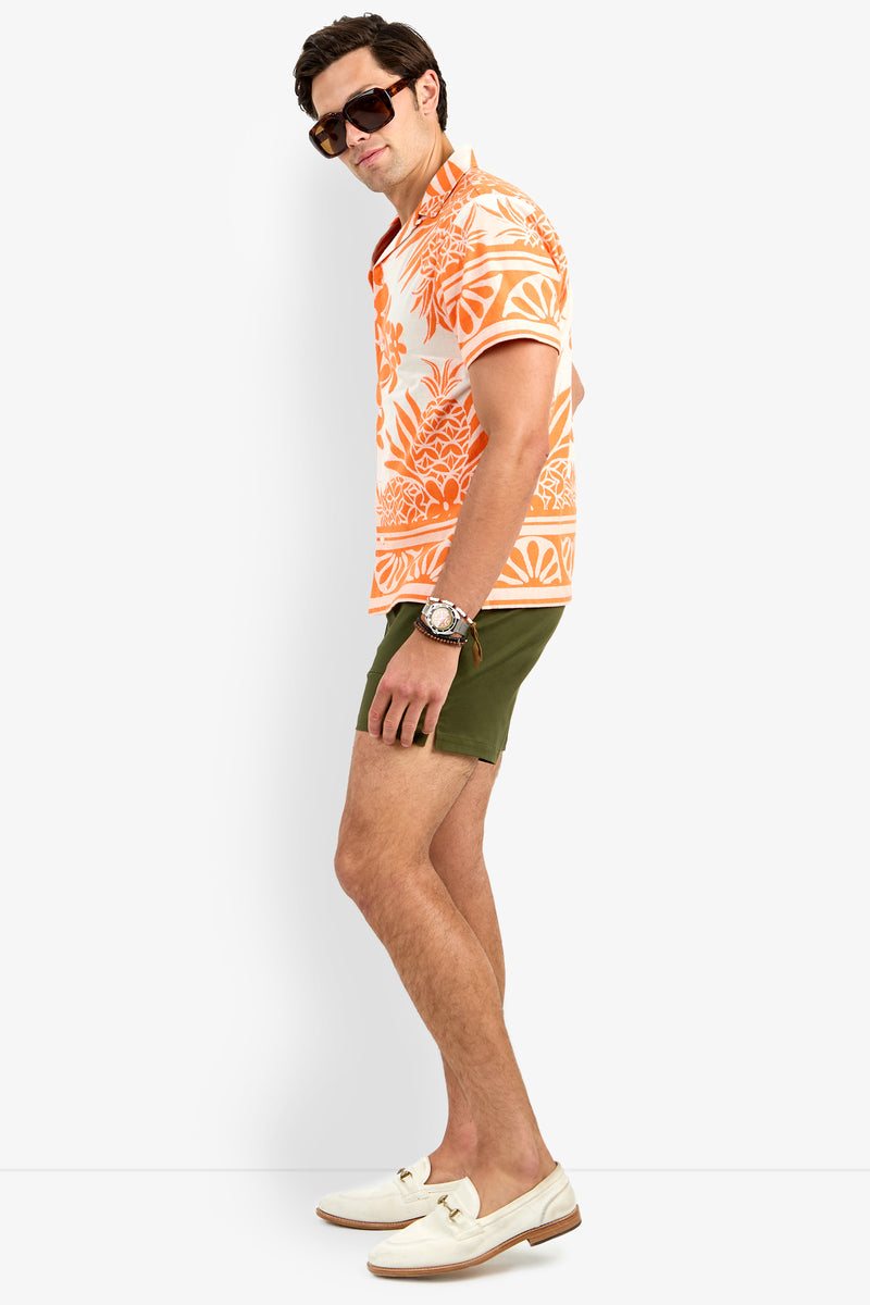 A man poses sideways in the Felix Shirt, featuring an orange floral print, paired with olive green shorts, white loafers, sunglasses, and a watch against a plain white background.