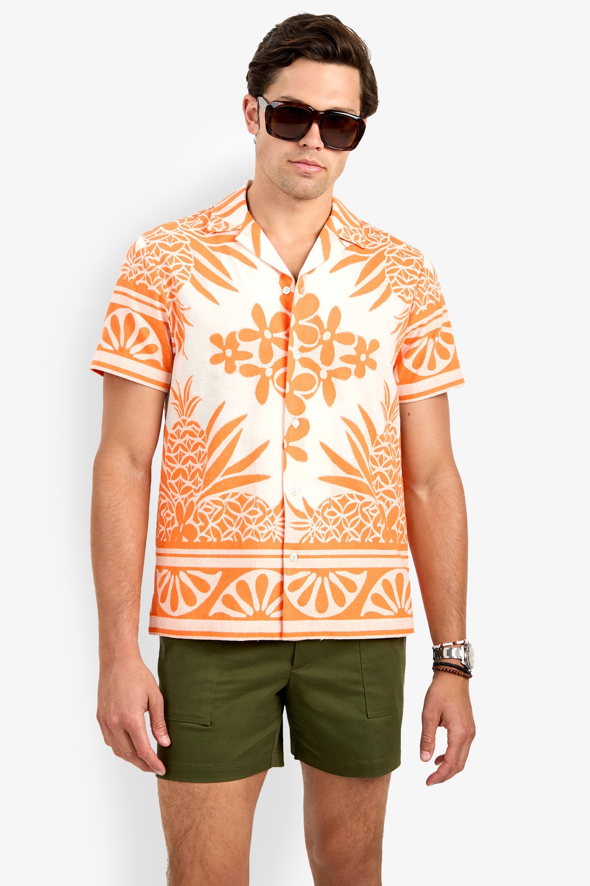 A man wearing dark sunglasses, the Felix Shirt—an orange and white Hawaiian shirt with floral and pineapple patterns—and olive green shorts stands against a plain white background.