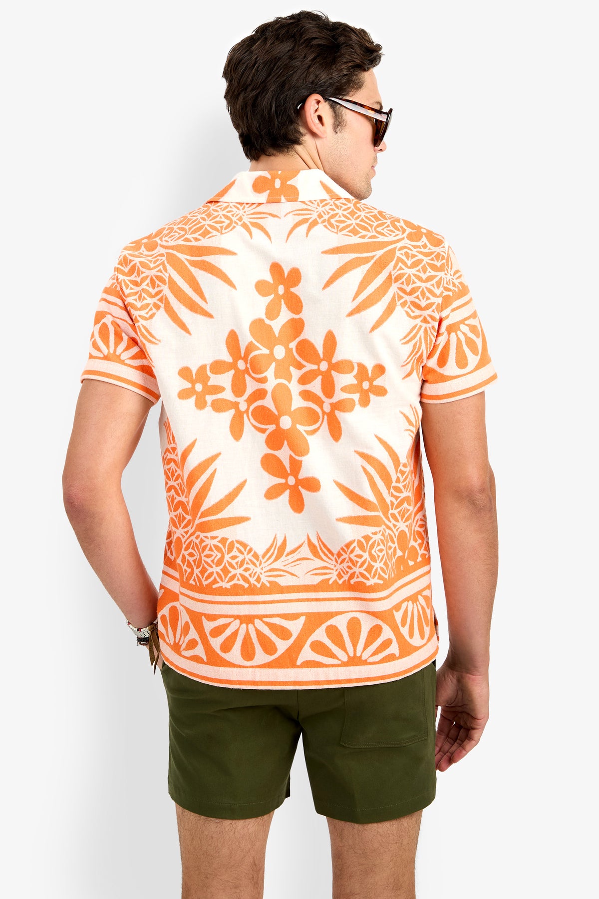 A man wearing the Felix Shirt, which features an orange and white floral pattern and short sleeves, paired with olive green shorts and sunglasses, is shown from the back against a plain light background.