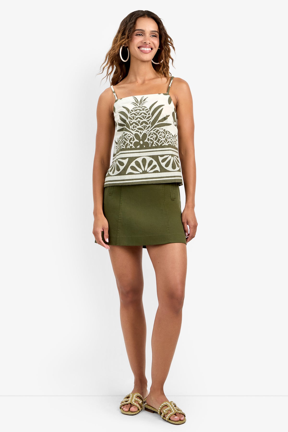 A woman smiles while standing, wearing the Marita Top with a pineapple and leaf design, an olive green mini skirt, gold hoop earrings, and gold sandals.