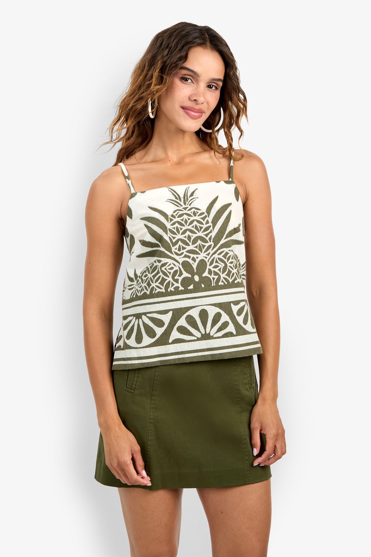 A woman wearing the Marita Top, a sleeveless blouse with a green pineapple pattern, paired with an olive green skirt, stands against a white background and smiles softly.