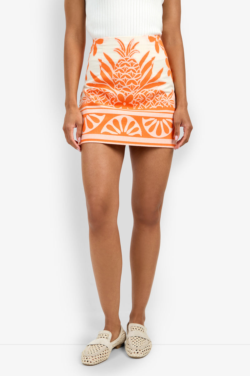 A person wearing a white sleeveless top, the High Rise Rico Skirt in orange and cream with a pineapple and geometric pattern, and cream woven slip-on shoes stands against a white background.
