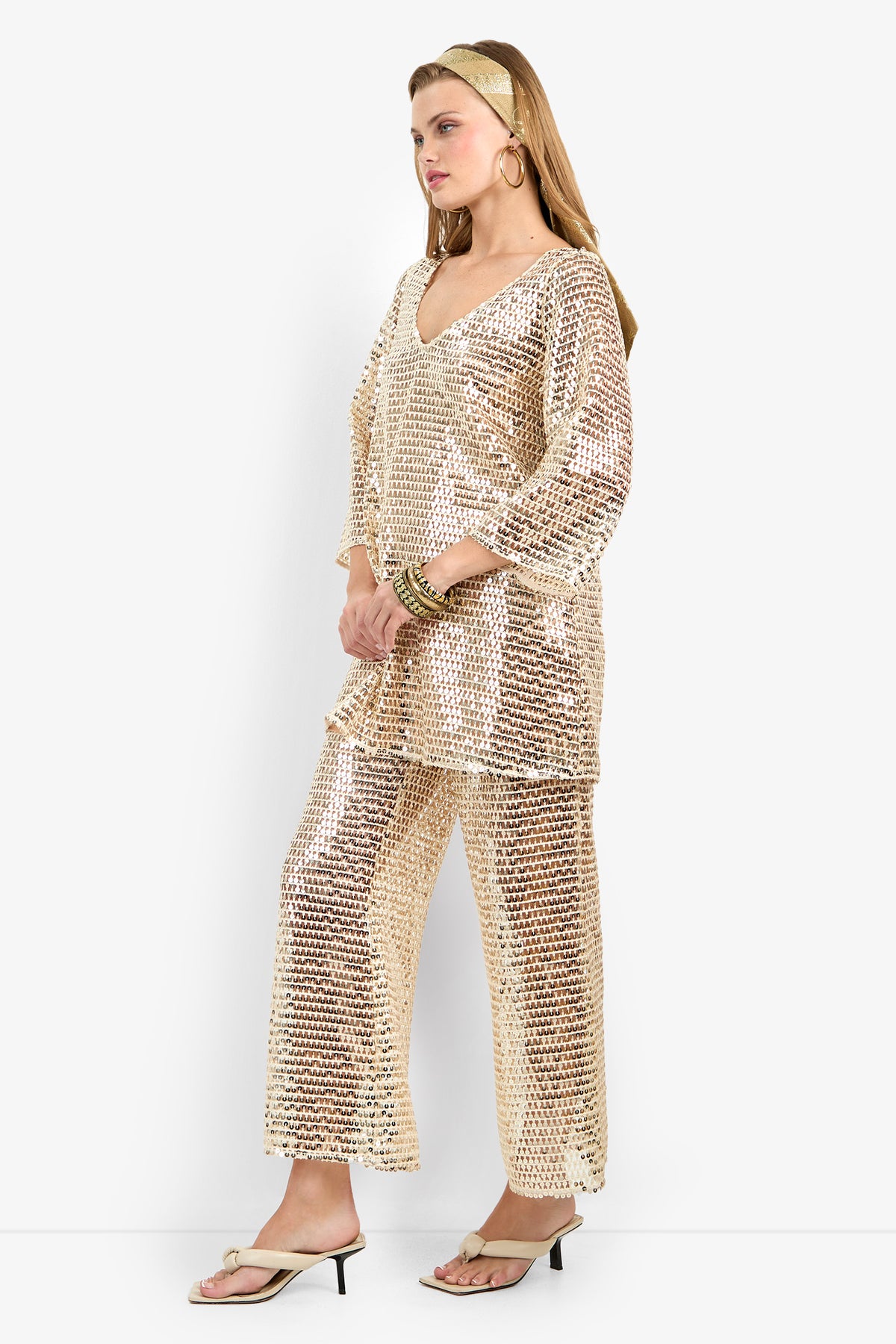 Woman stands in profile wearing the Guava Dress, a loose-fitting gold sequin outfit with wide sleeves. She completes her look with gold hoop earrings, bangles, a headband, and open-toe heeled sandals.