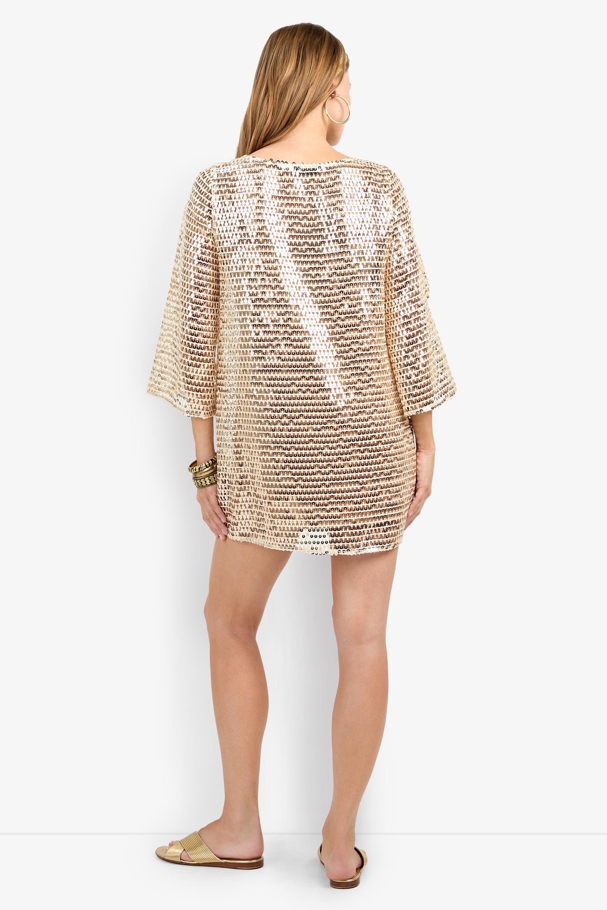 A woman with long light brown hair faces away, wearing the Guava Dress—a loose gold sequin-embellished tunic with wide sleeves. She accessorizes with flat sandals and gold bangles against a plain white background.