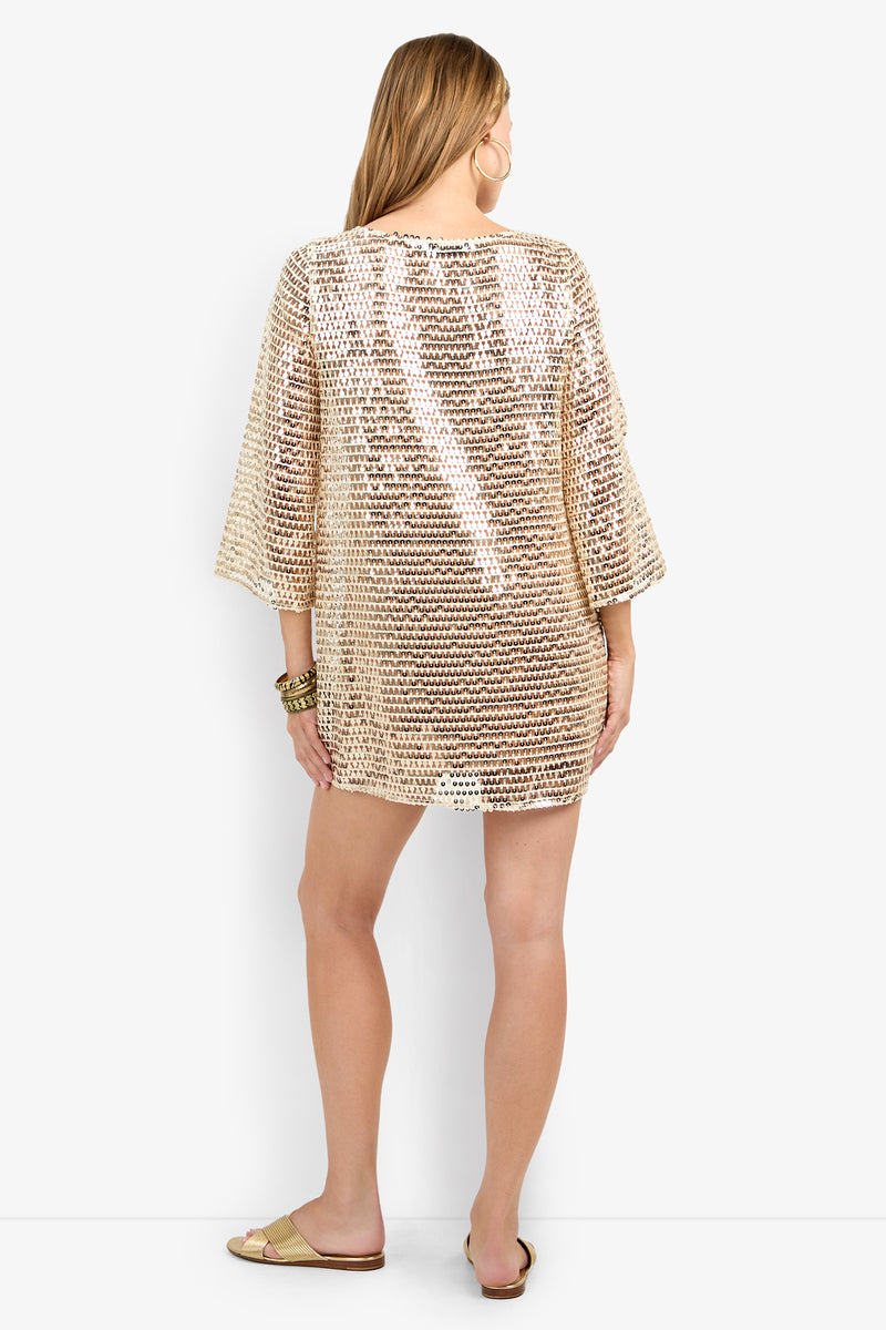 A woman with long light brown hair faces away, wearing the Guava Dress—a loose gold sequin-embellished tunic with wide sleeves. She accessorizes with flat sandals and gold bangles against a plain white background.