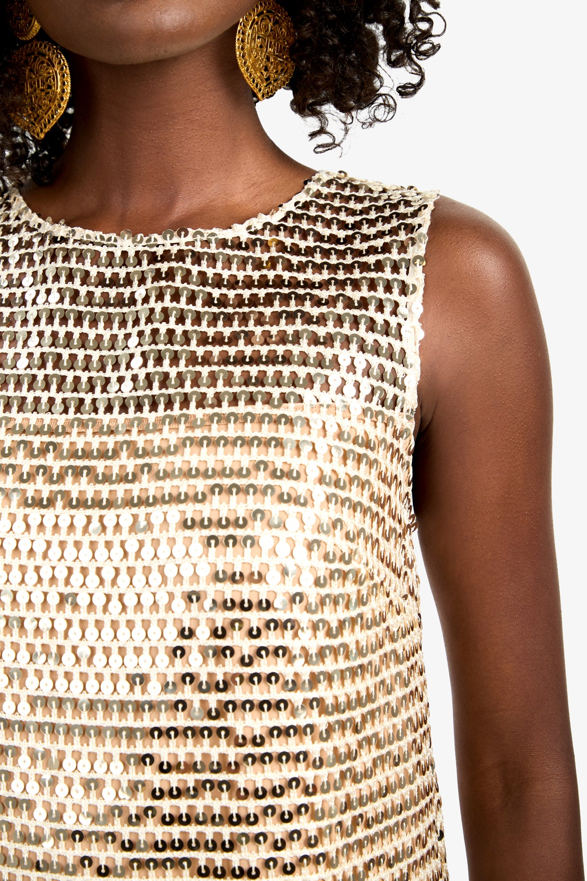 A woman models the Eleanor Dress, a sleeveless, gold sequin-covered design. Large gold earrings accent her look. Only her lower face and upper torso are shown against a white background.