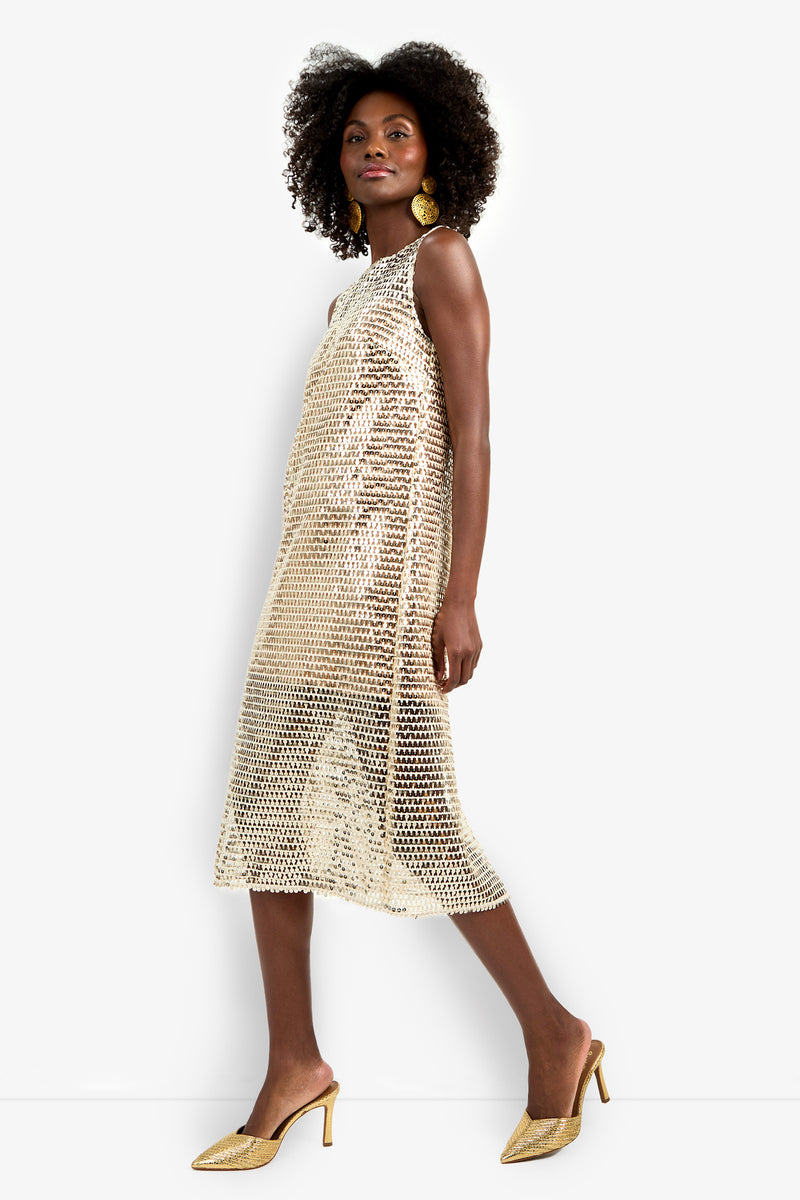 A woman poses confidently in profile against a white background, wearing the Eleanor Dress—a sleeveless, knee-length gold sequin dress with a loose fit—styled with hoop earrings, gold heels, and natural curly hair.