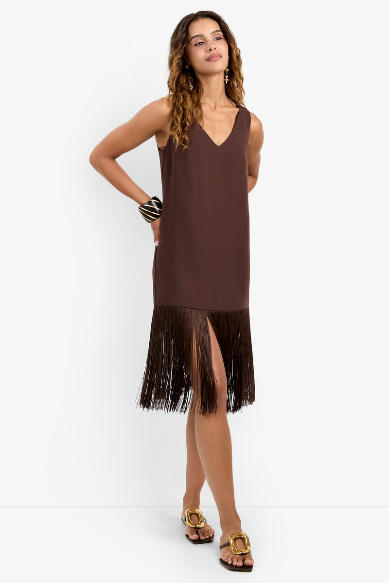 A woman stands against a white background wearing the AYMANI DRESS in Carob—a sleeveless, durable polyester dress with a V-neck and long fringe at the hem—accessorized with a wide bracelet and brown sandals.