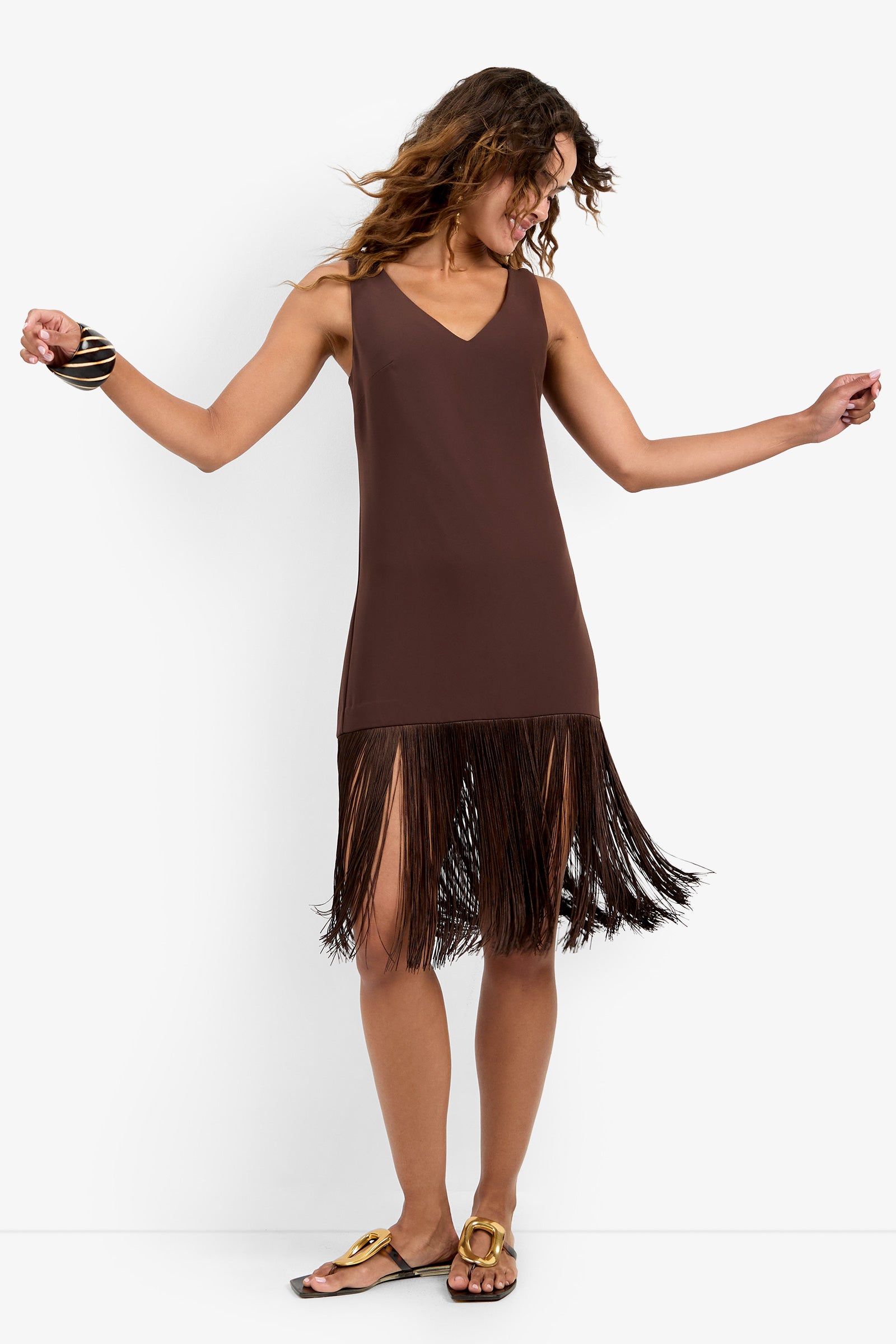 A woman in the AYMANI DRESS—a sleeveless brown polyester dress with fringe at the hem—dances, smiling with her head turned sideways. She has wavy hair, wears sandals, and a chunky bracelet on her left wrist.