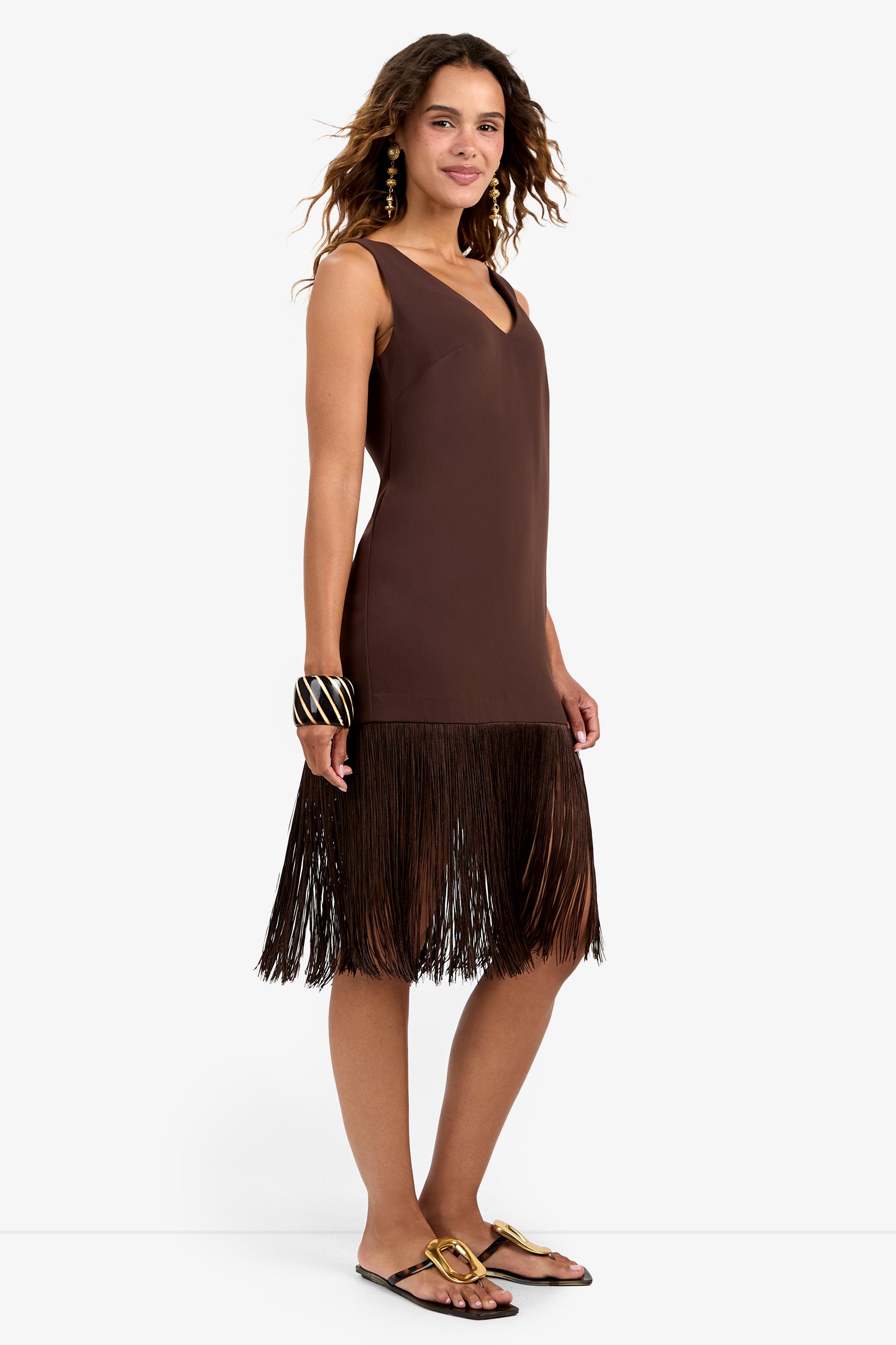 A woman in the sleeveless brown Aymani Dress with a V-neckline and fringe hem, gold earrings, chunky bracelet, and gold sandals stands against a plain white background, smiling softly.