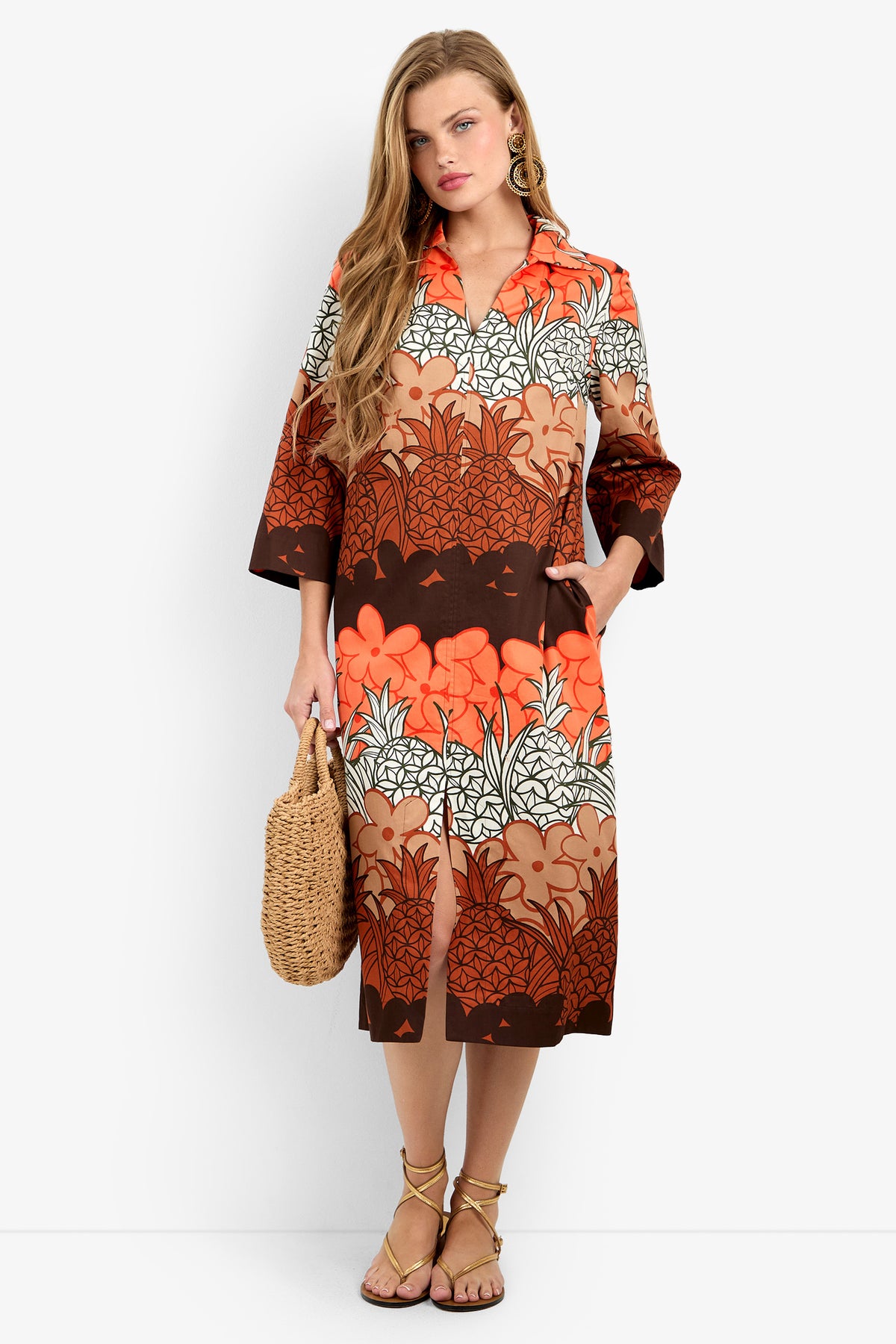 A woman wears the KAMIA DRESS in Meli Kalima, a knee-length, long-sleeve 100% cotton dress with an orange, brown, and white floral print. She stands against a white background holding a woven handbag and wearing strappy sandals.