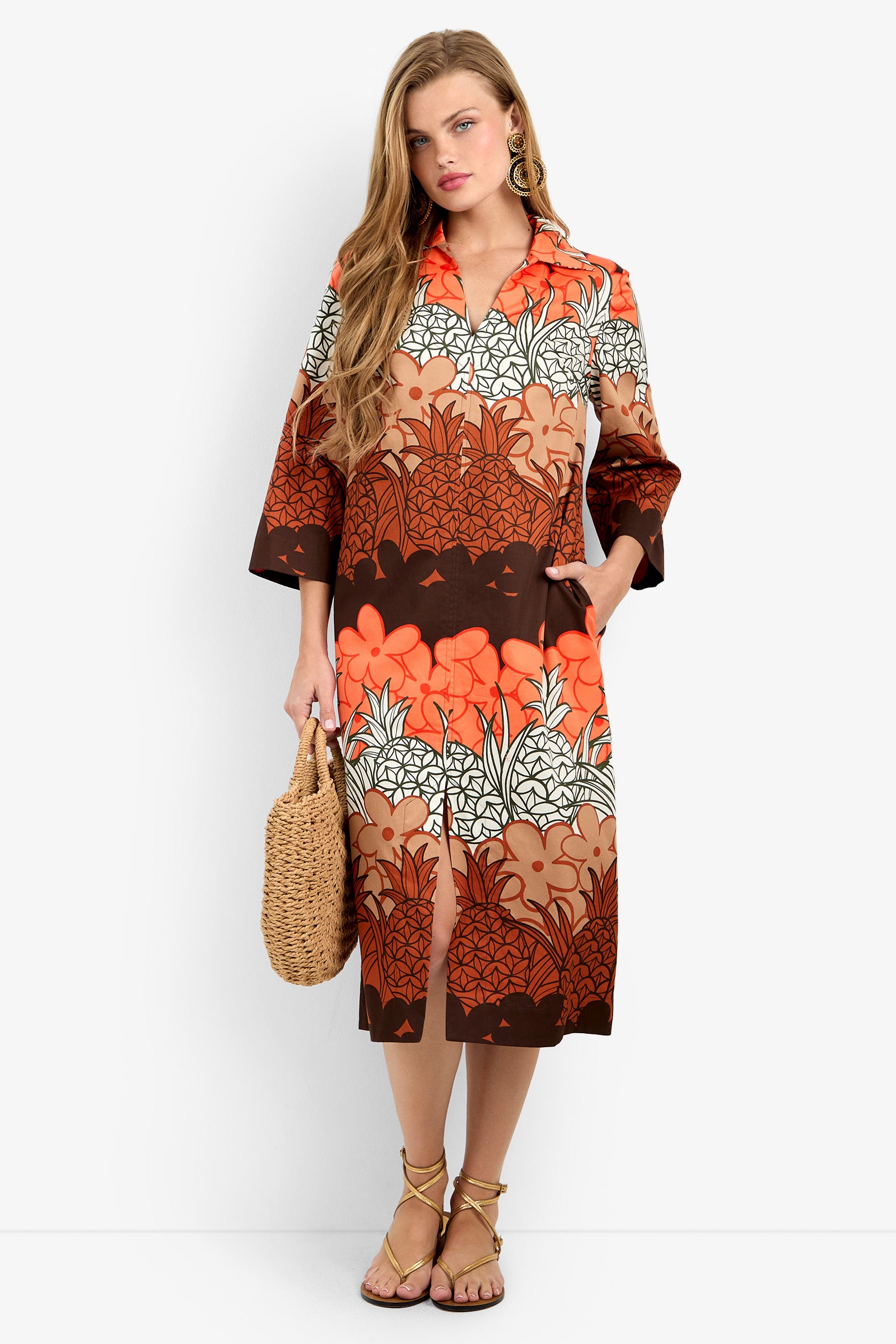 A woman wears the KAMIA DRESS in Meli Kalima, a knee-length, long-sleeve 100% cotton dress with an orange, brown, and white floral print. She stands against a white background holding a woven handbag and wearing strappy sandals.