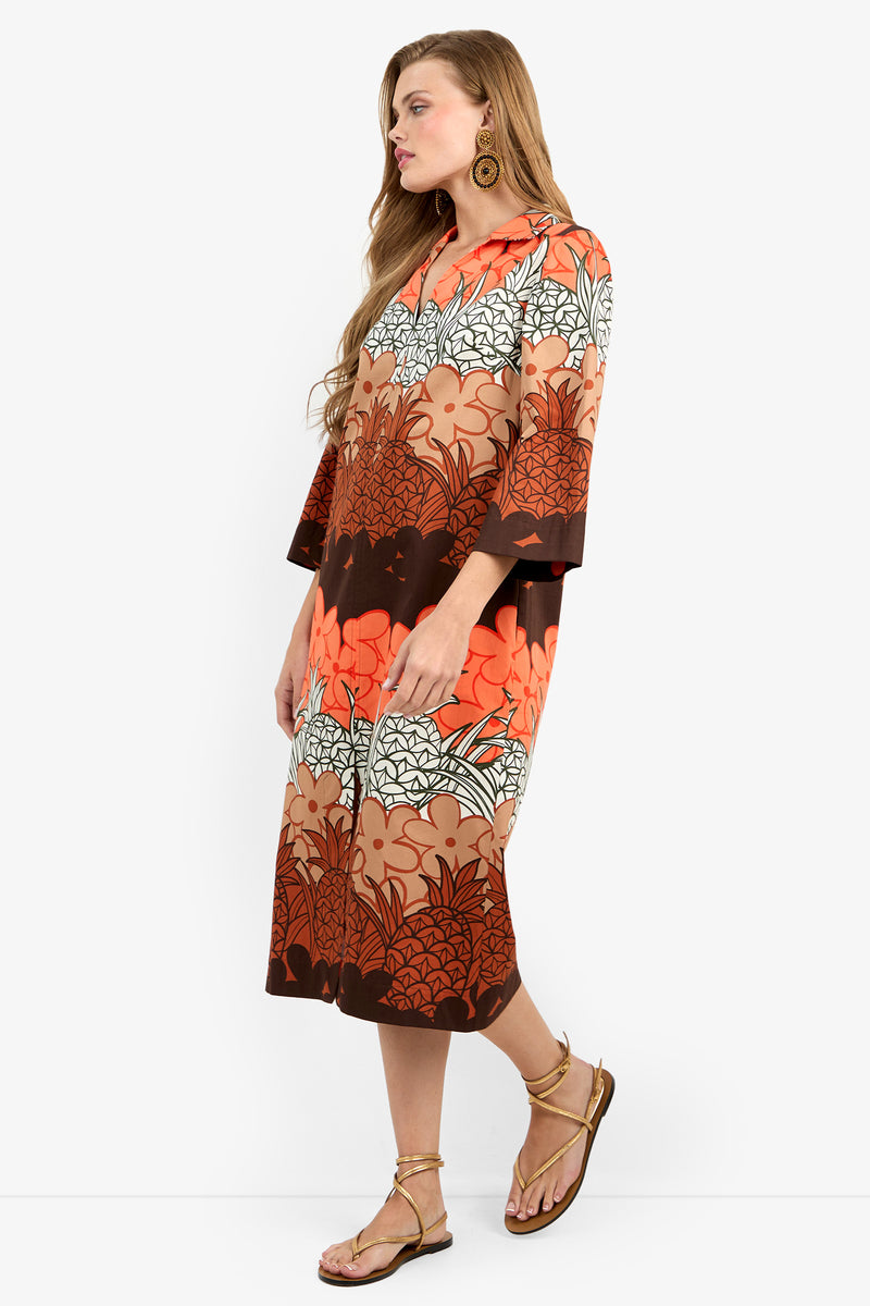 A woman stands sideways wearing the Kamia Dress—a loose, knee-length style with a bold brown, orange, and white floral print—paired with flat sandals and large round earrings.