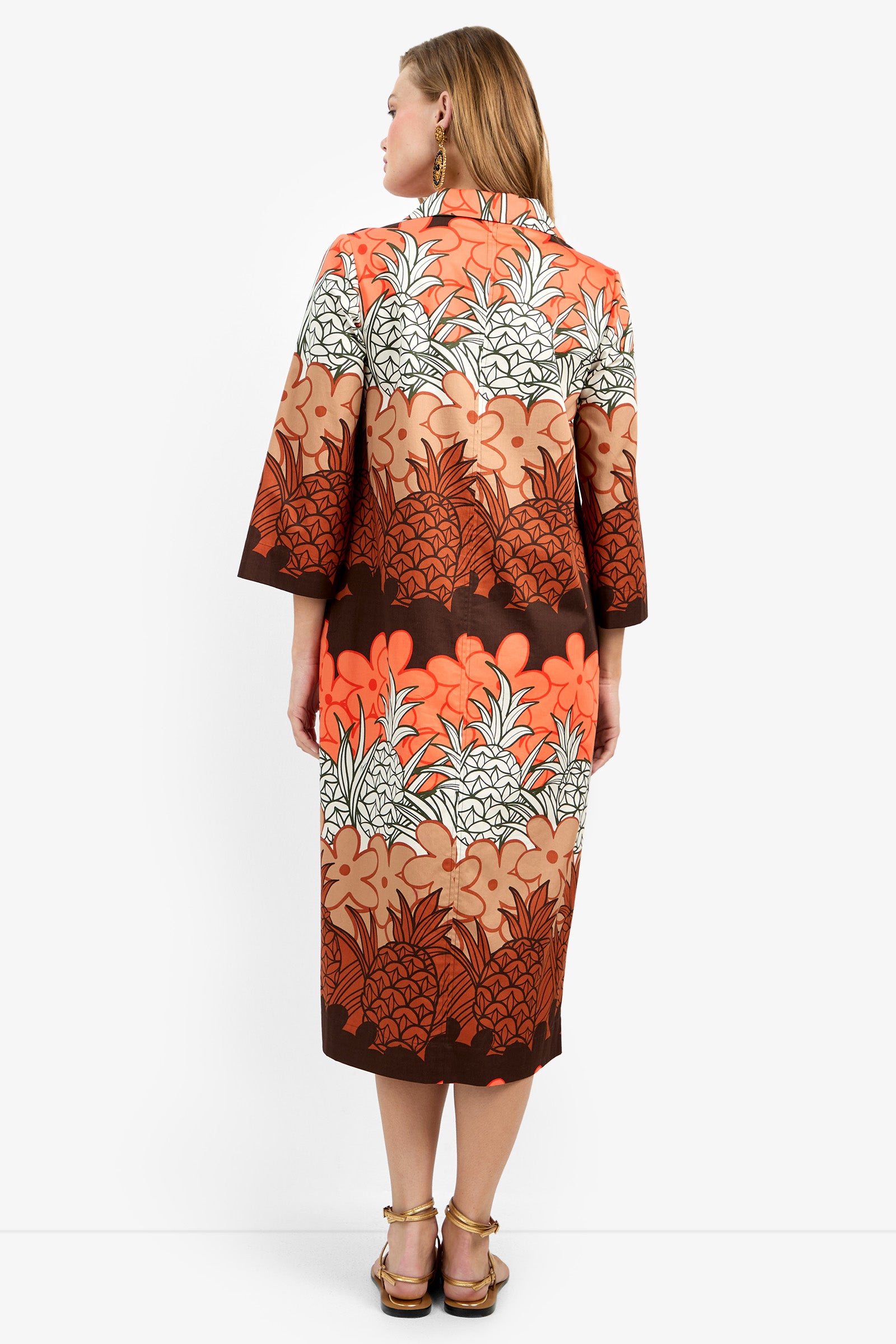 A woman with long, straight hair wears the Kamia Dress—a knee-length brown-orange floral dress with three-quarter sleeves—paired with sandals and earrings against a plain white background.