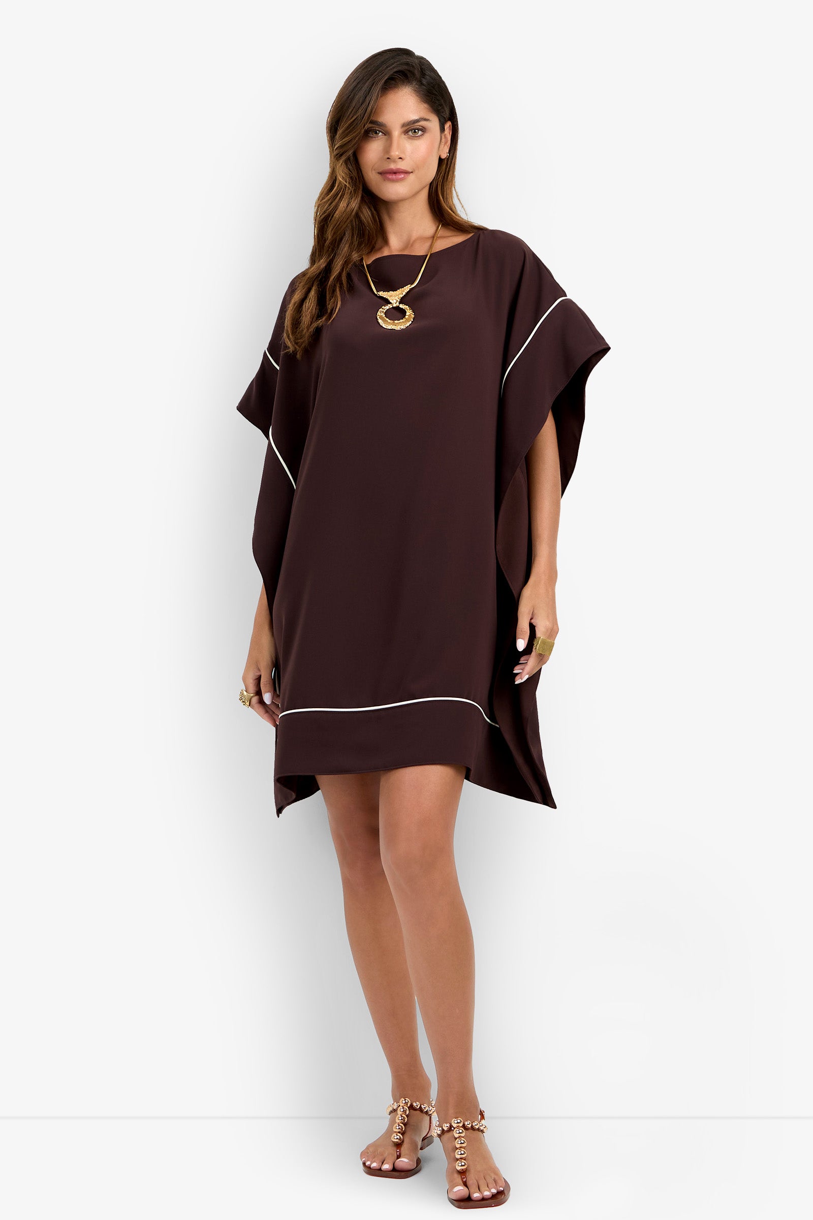 A woman wearing the Kalena Caftan Dress—a loose, dark brown short dress with white trim—paired with a gold necklace, rings, and sandals, stands against a plain white background.