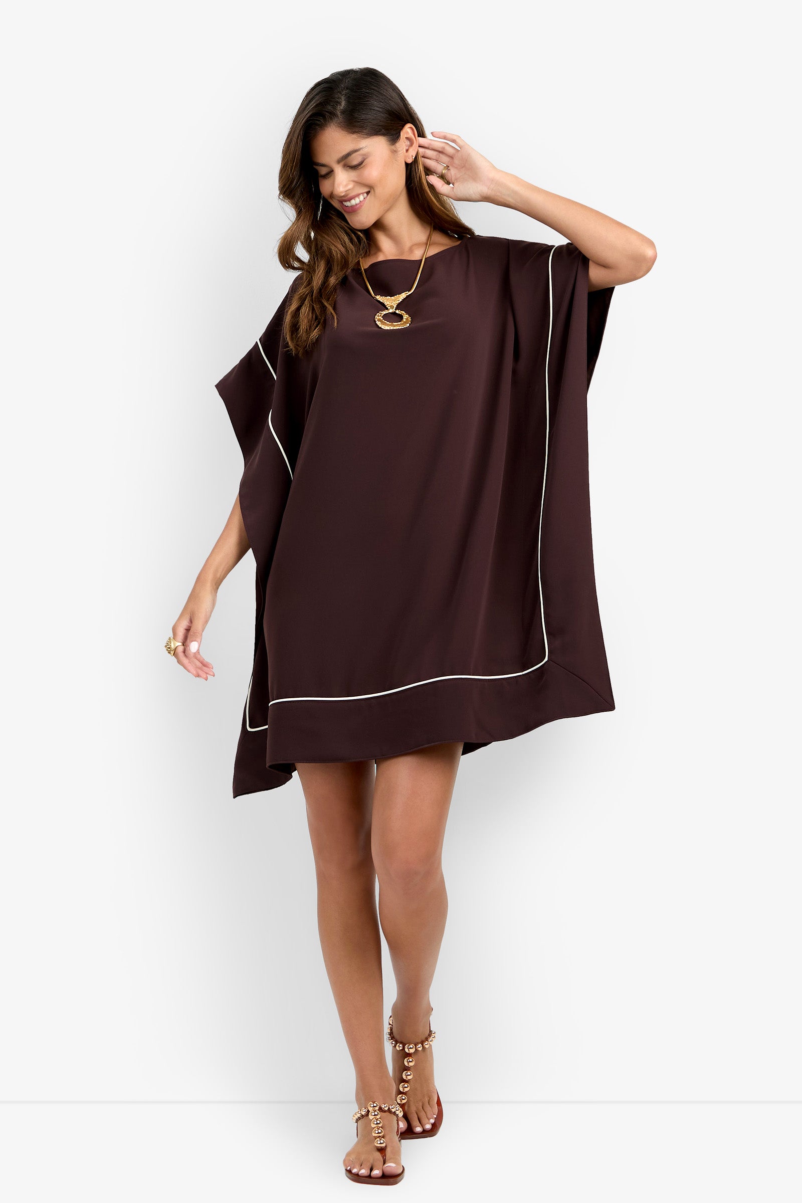 A woman is smiling and looking down while standing against a plain white background, wearing the Kalena Caftan Dress in loose, dark brown fabric with white trim, paired with a gold necklace, ring, and sandals.