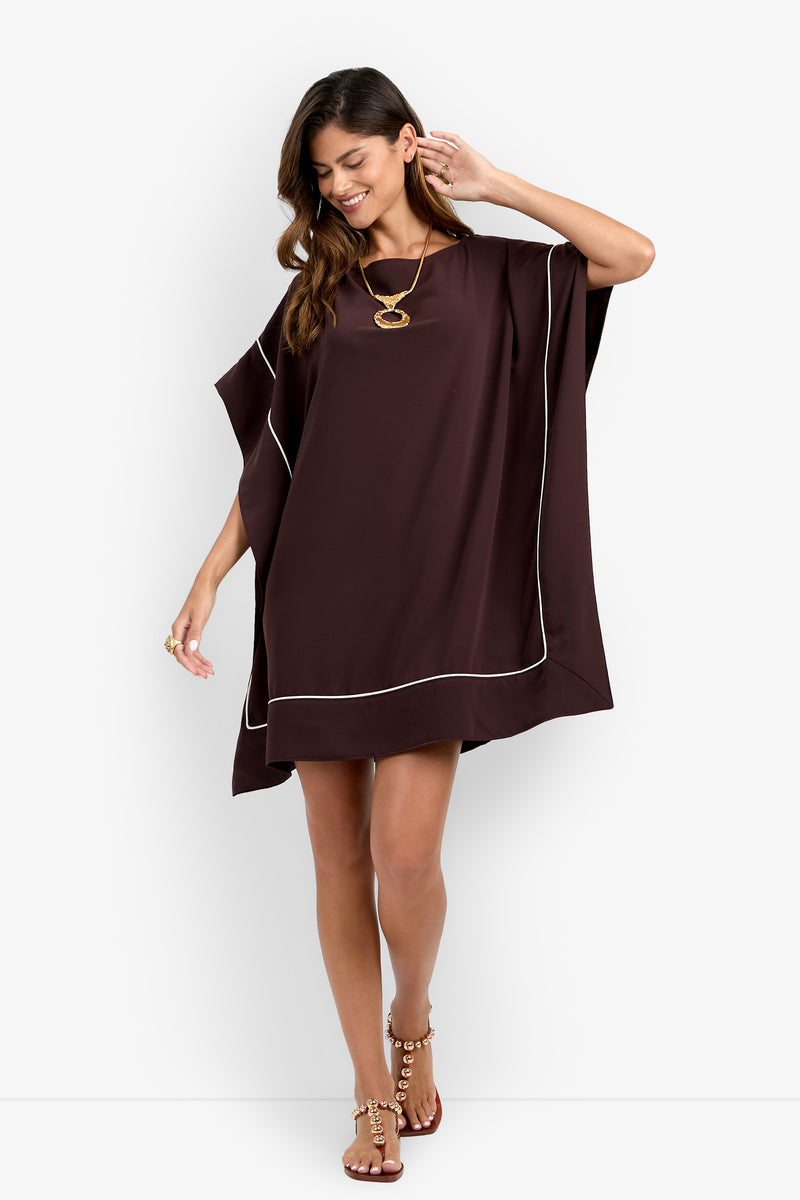 A woman is smiling and looking down while standing against a plain white background, wearing the Kalena Caftan Dress in loose, dark brown fabric with white trim, paired with a gold necklace, ring, and sandals.