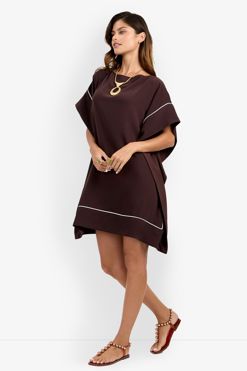 A woman wearing the Kalena Caftan Dress—a loose-fitting, short brown dress with white trim—paired with a gold necklace, rings, and brown flat sandals adorned with pearls, stands against a plain white background.