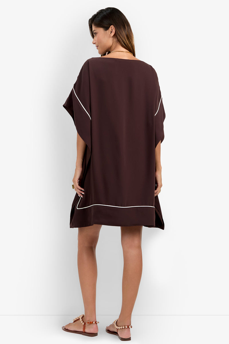 A woman stands with her back to the camera, wearing the Kalena Caftan Dress—a loose, dark brown dress with white piping, wide short sleeves, and a hem that falls above her knees. She pairs it with flat sandals against a plain white background.