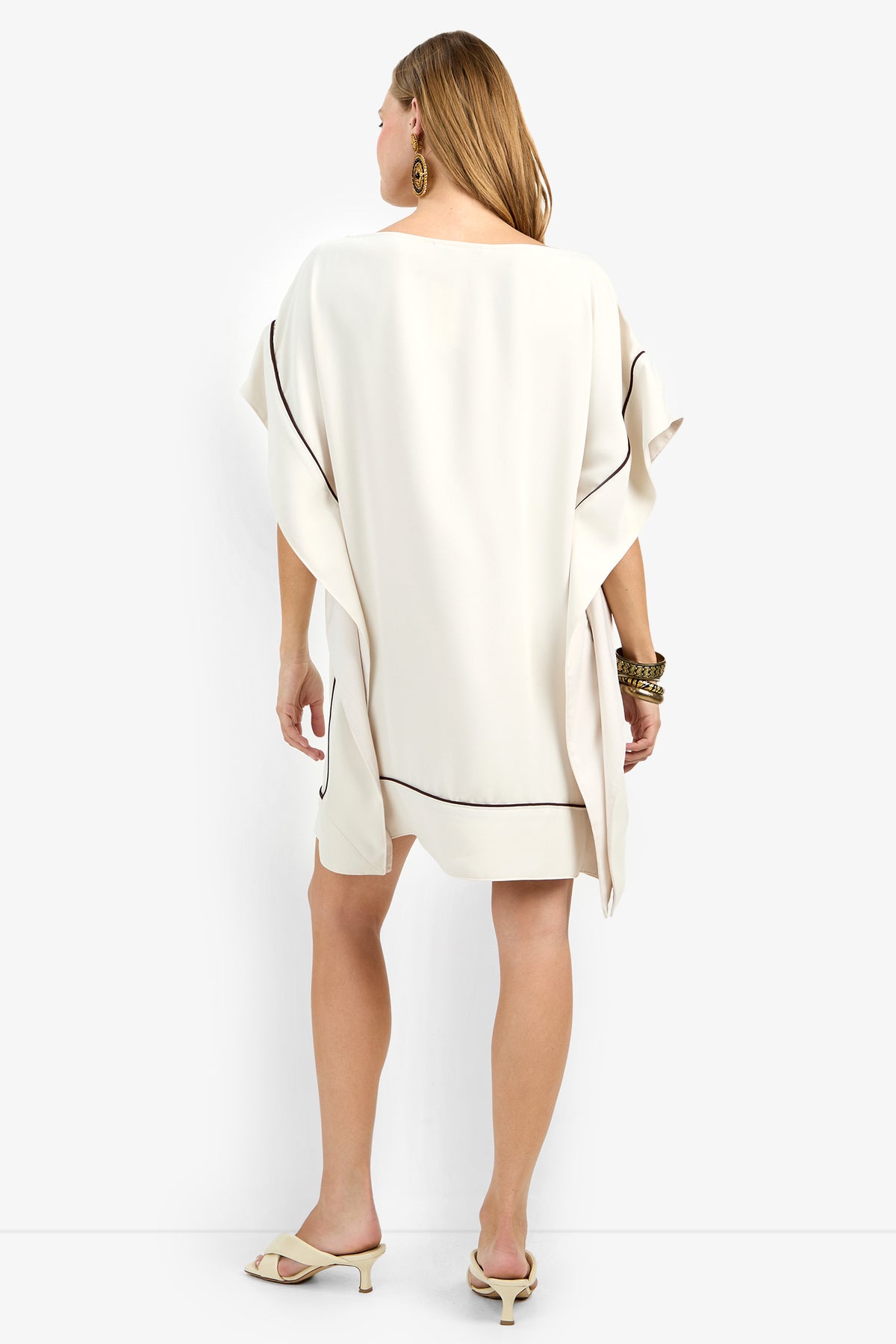 A woman faces away in the Kalena Dress—loose, cream with black piping—paired with beige heeled sandals, gold hoop earrings, and bracelets. Her light brown hair is down against a plain white background.