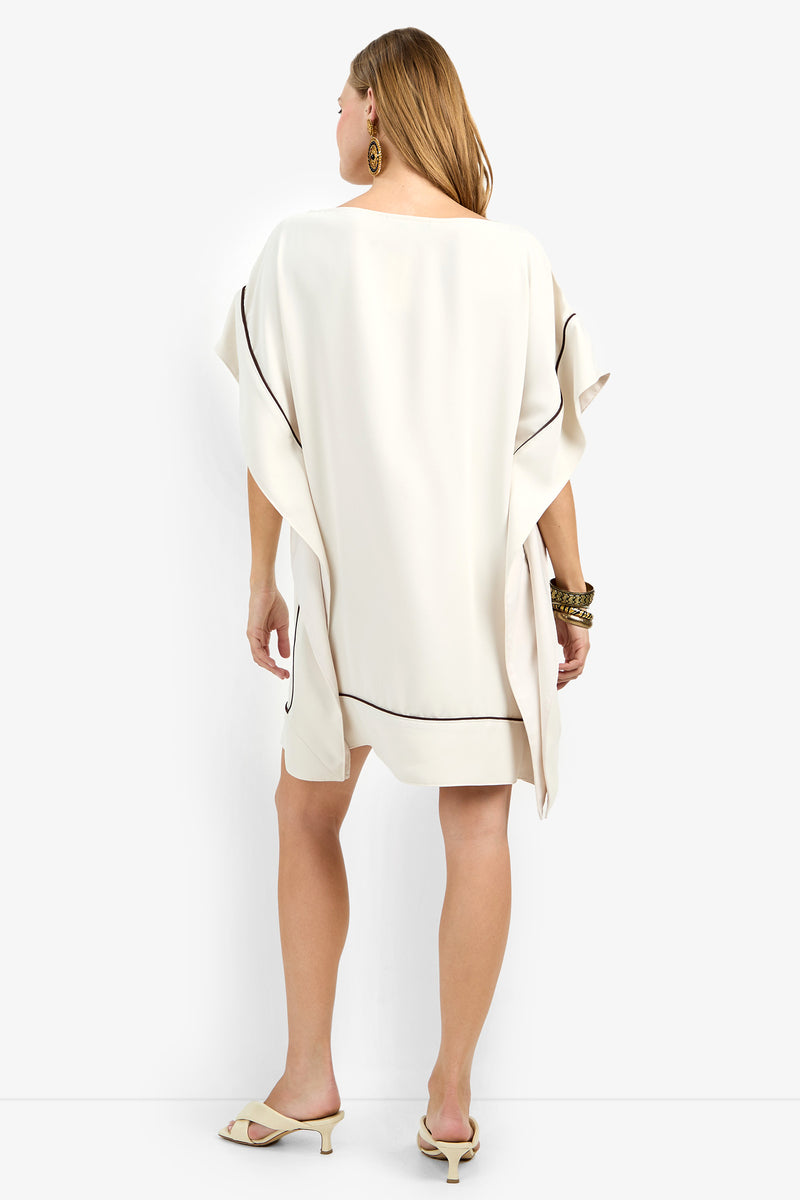 A woman faces away in the Kalena Dress—loose, cream with black piping—paired with beige heeled sandals, gold hoop earrings, and bracelets. Her light brown hair is down against a plain white background.