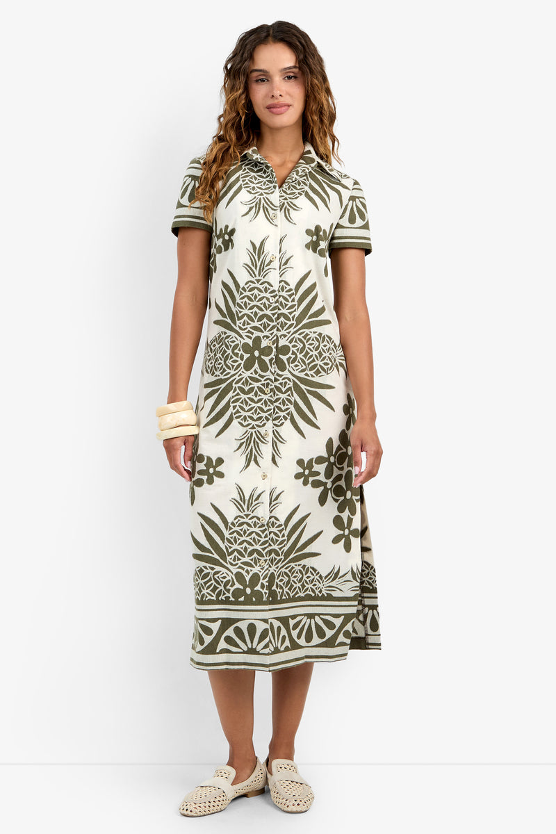 A woman in the Abba Dress—a cream, short-sleeve collared midi with green tropical pineapple and leaf prints—paired with beige flat woven shoes and chunky bangles, stands against a white background.