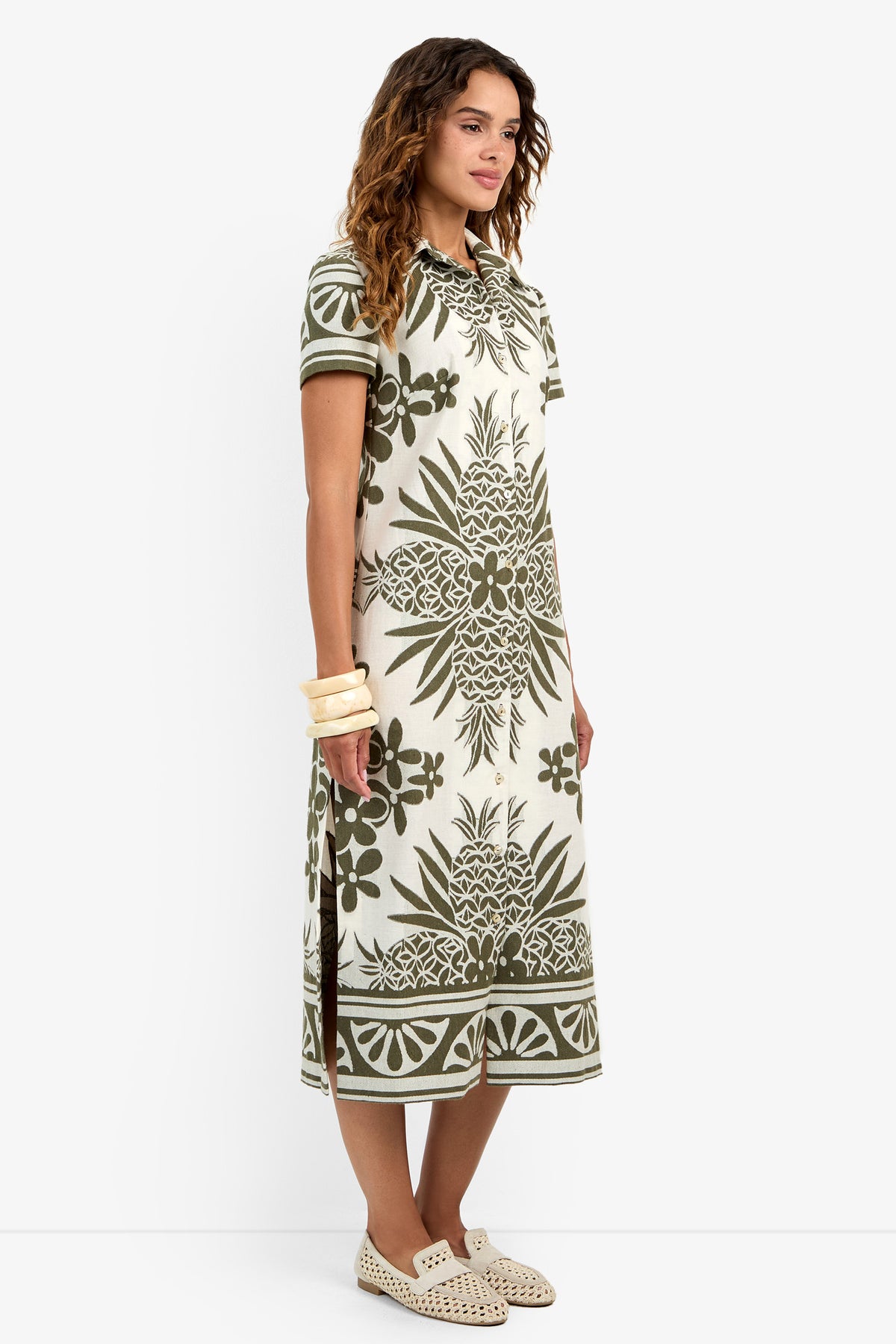 A woman with wavy hair and a calm expression stands in a side pose wearing the Abba Dress—a cream midi dress with green floral and geometric prints—paired with cream slip-on shoes and multiple bangle bracelets.
