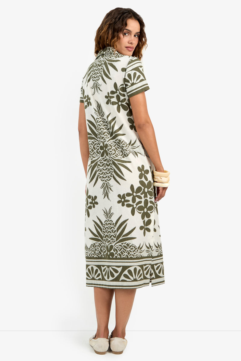 A woman stands facing away, wearing the Abba Dress— a short-sleeved, knee-length dress featuring green and white Polynesian-inspired patterns—paired with bracelets and flat shoes.