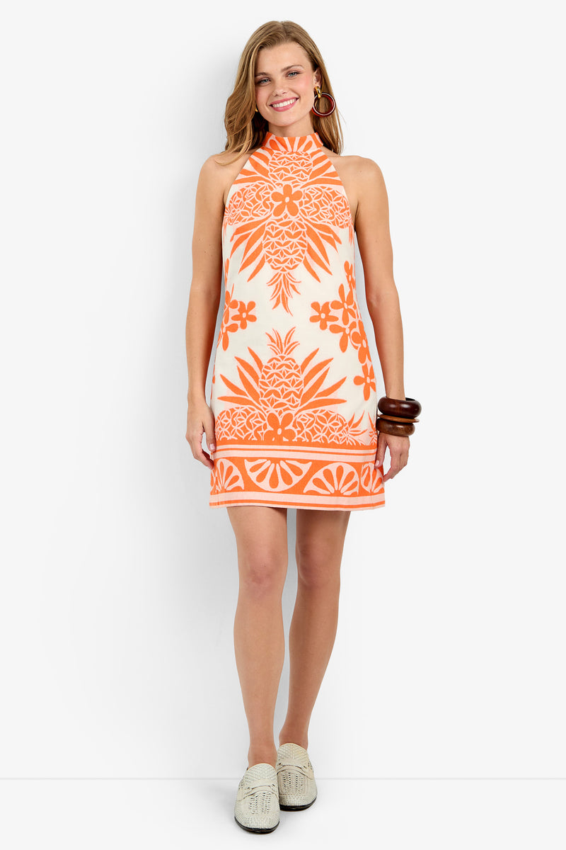 Smiling, a woman wears the GARAM DRESS in ORANGE OASIS/SANCERRE, a sleeveless, short white dress with bold orange floral and pineapple patterns. She accessorizes with large hoops, chunky brown bracelets, and white slip-on shoes.