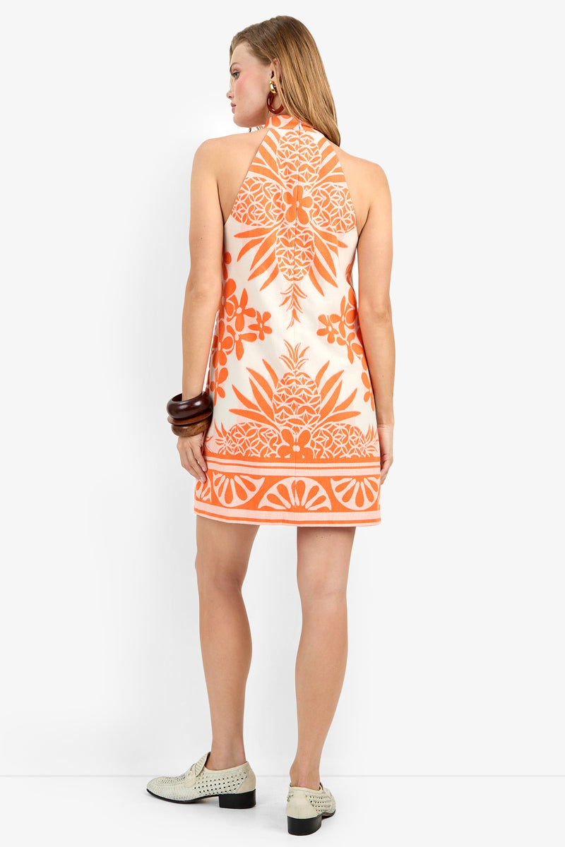 A woman with light brown hair worn down faces away, wearing the Garam Dress—a sleeveless, halter-neck mini dress with an orange and white tropical print—paired with cream shoes and statement bangles.
