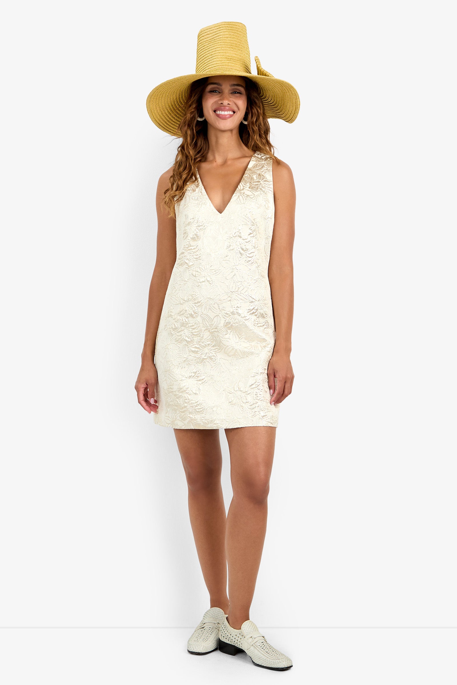 A woman stands smiling against a plain white background, wearing the Addy Dress—a sleeveless cream-colored V-neck dress—paired with a wide-brimmed straw hat and white textured slip-on shoes.