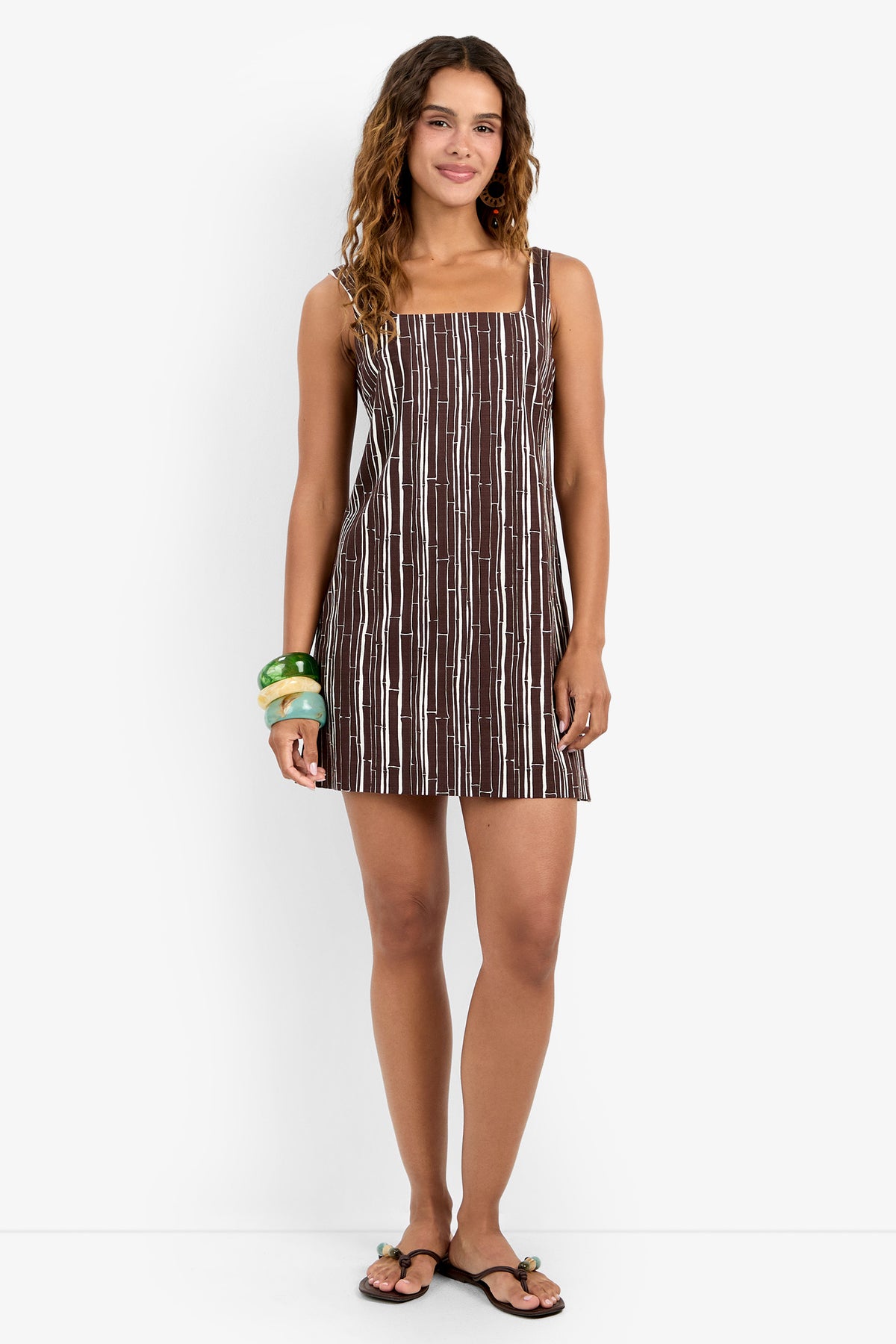 A woman with wavy hair wears the ASPIRE DRESS, a brown and white striped sleeveless mini made from soft viscose, paired with black sandals and chunky green and blue bracelets, standing against a plain white background.
