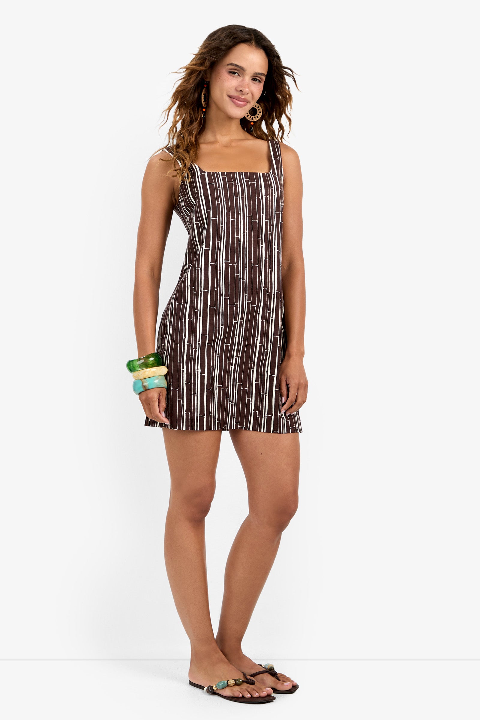 A woman with wavy hair and a slight smile wears the Aspire Dress—brown and white striped, sleeveless, mini length—paired with large green bangles, hoop earrings, and sandals against a white background.