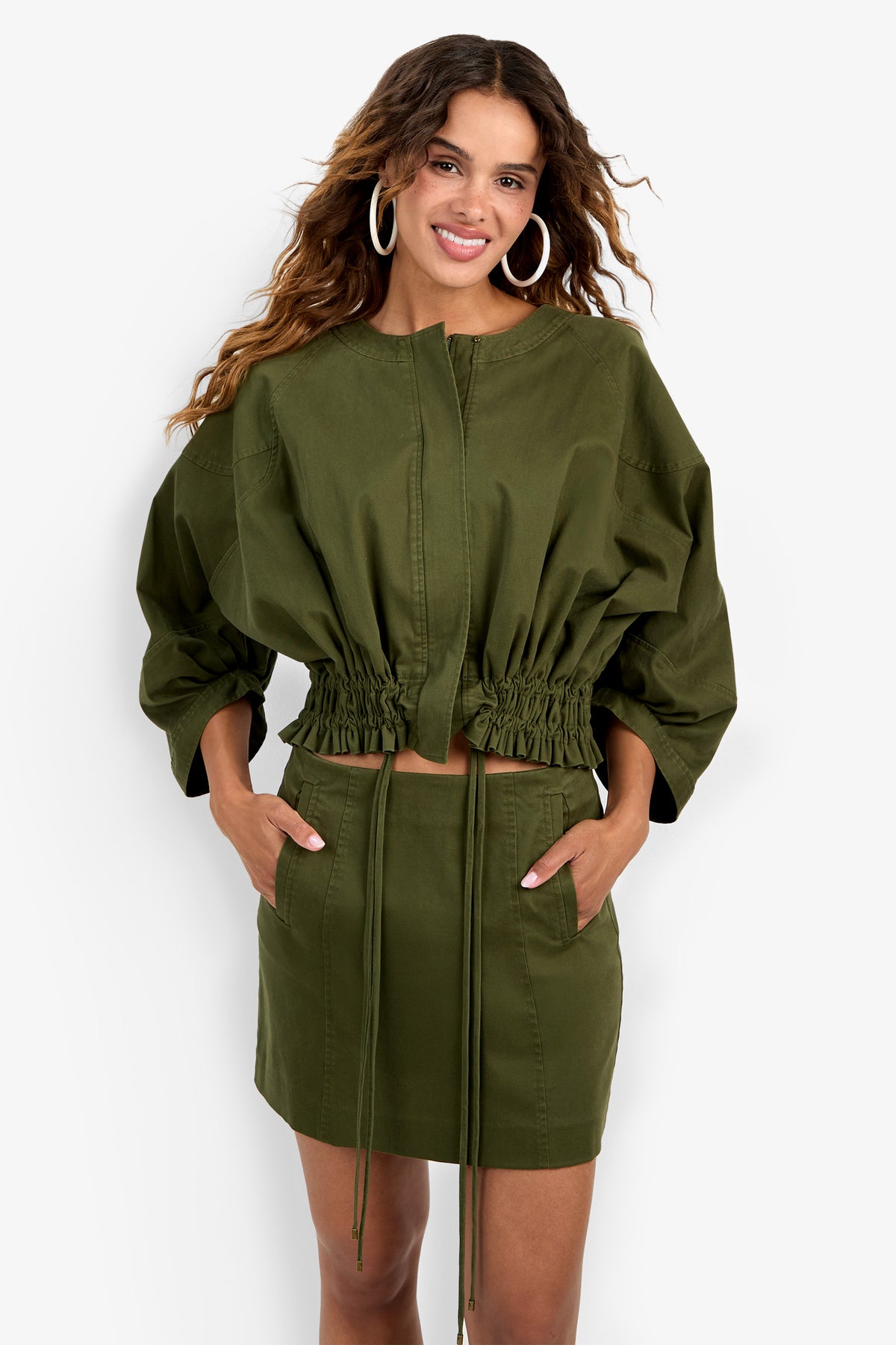 A woman wearing the Prita Jacket in olive green, styled with a matching high-waisted mini skirt. She has long wavy hair, large hoop earrings, and her hands in her skirt pockets.